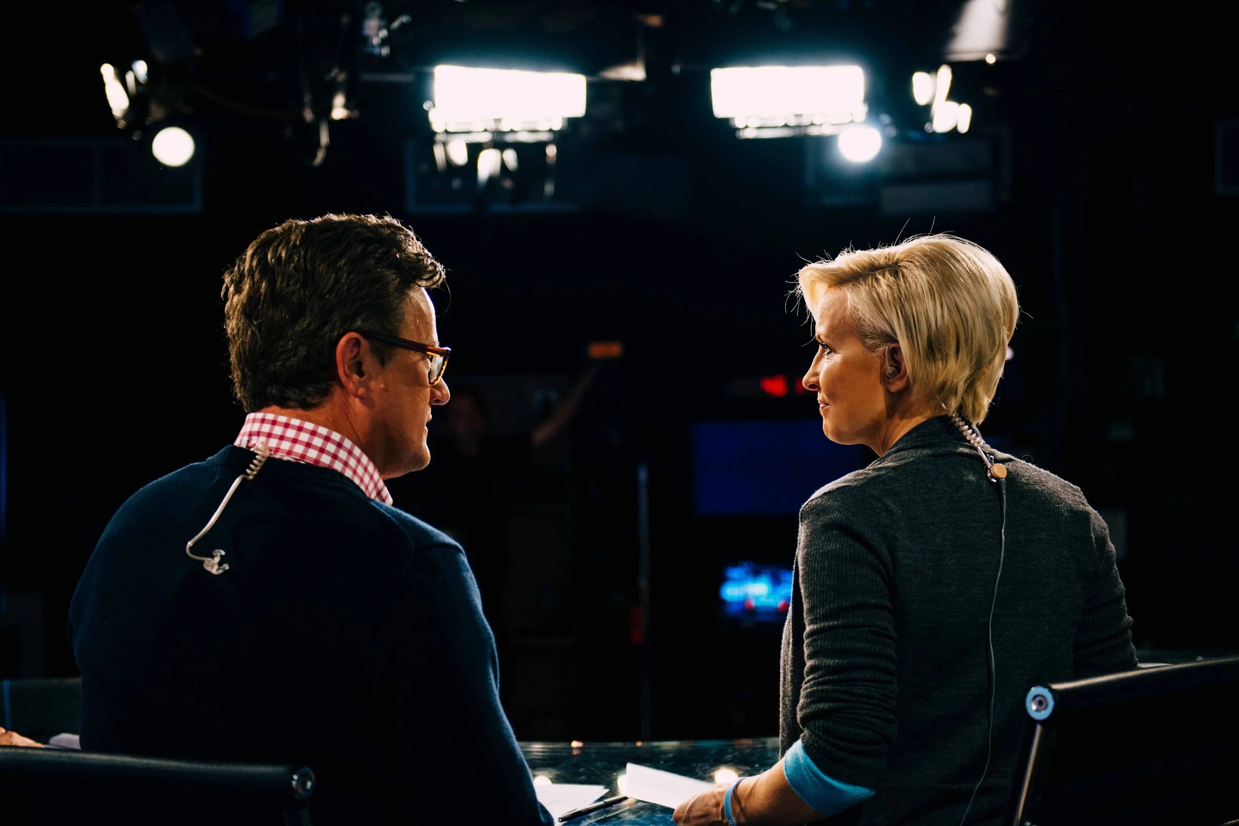 Two people, a man and a woman, sit facing each other in a television studio, engaged in conversation. Bright studio lights are above them.