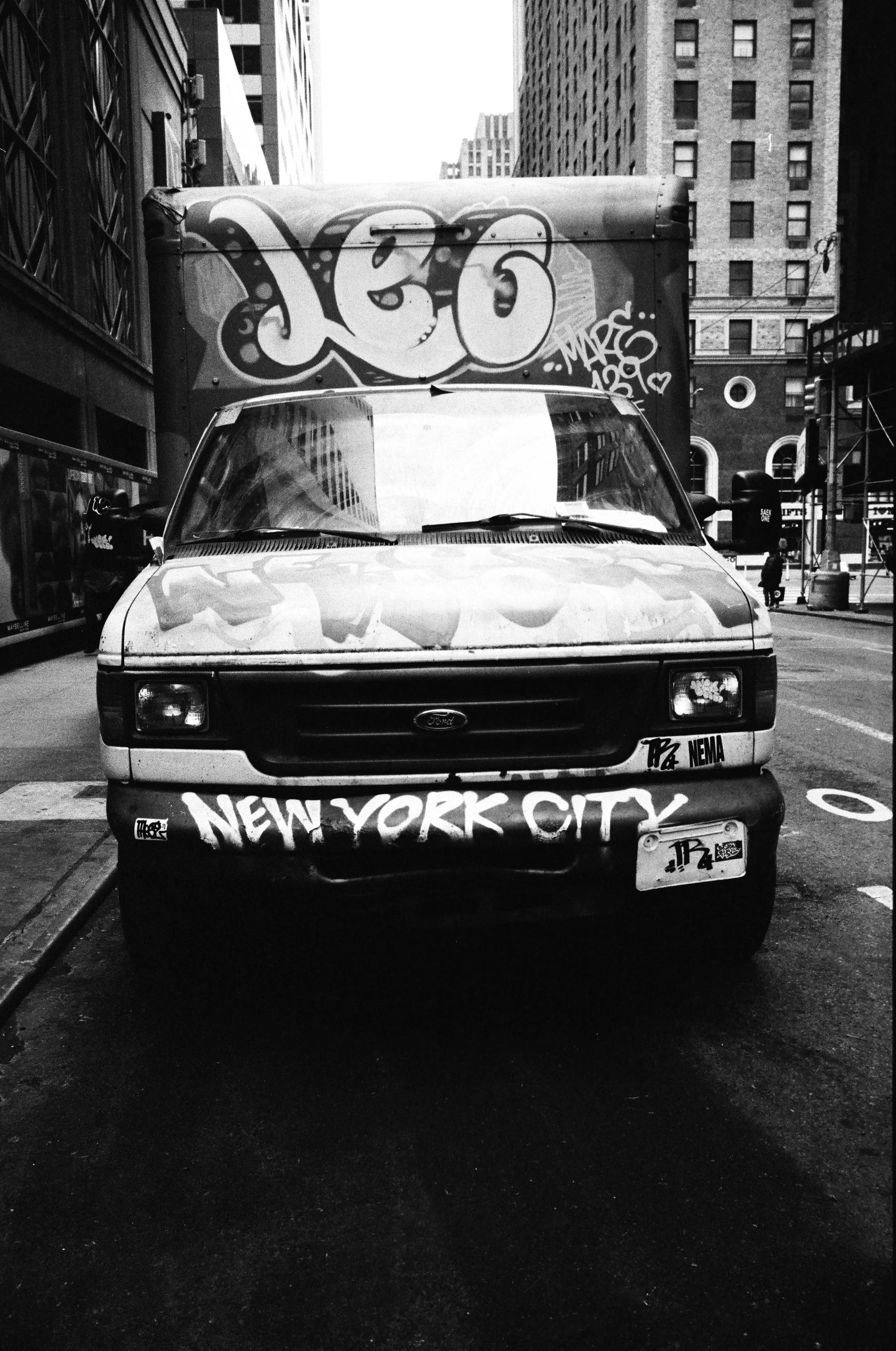 A graffiti-covered delivery truck parked on a city street in New York City, with buildings and pedestrians in the background, all in black and white.
