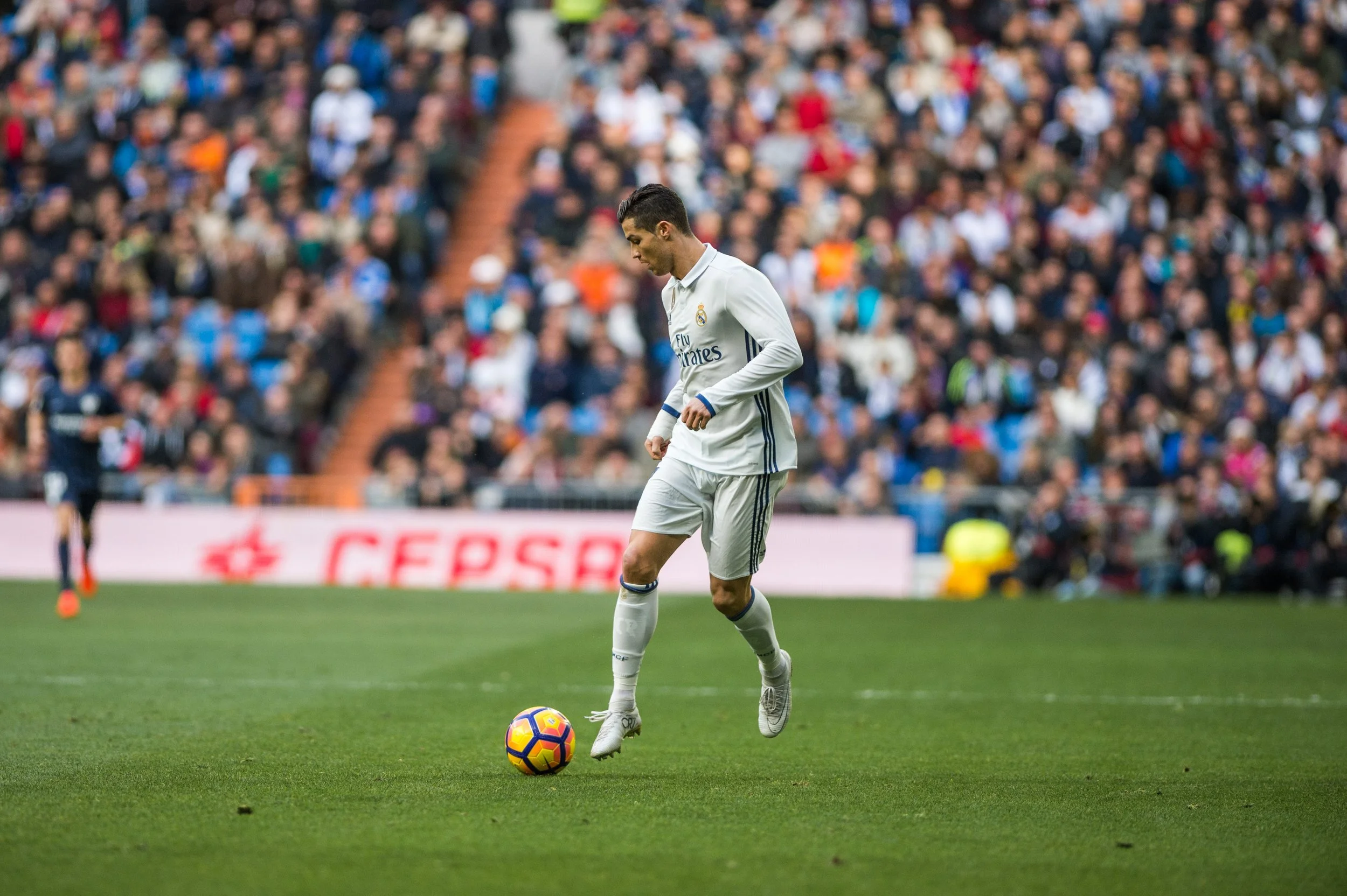 Soccer player in white uniform, standing on a green field, preparing to kick a yellow and purple soccer ball, with a large crowd in the background.