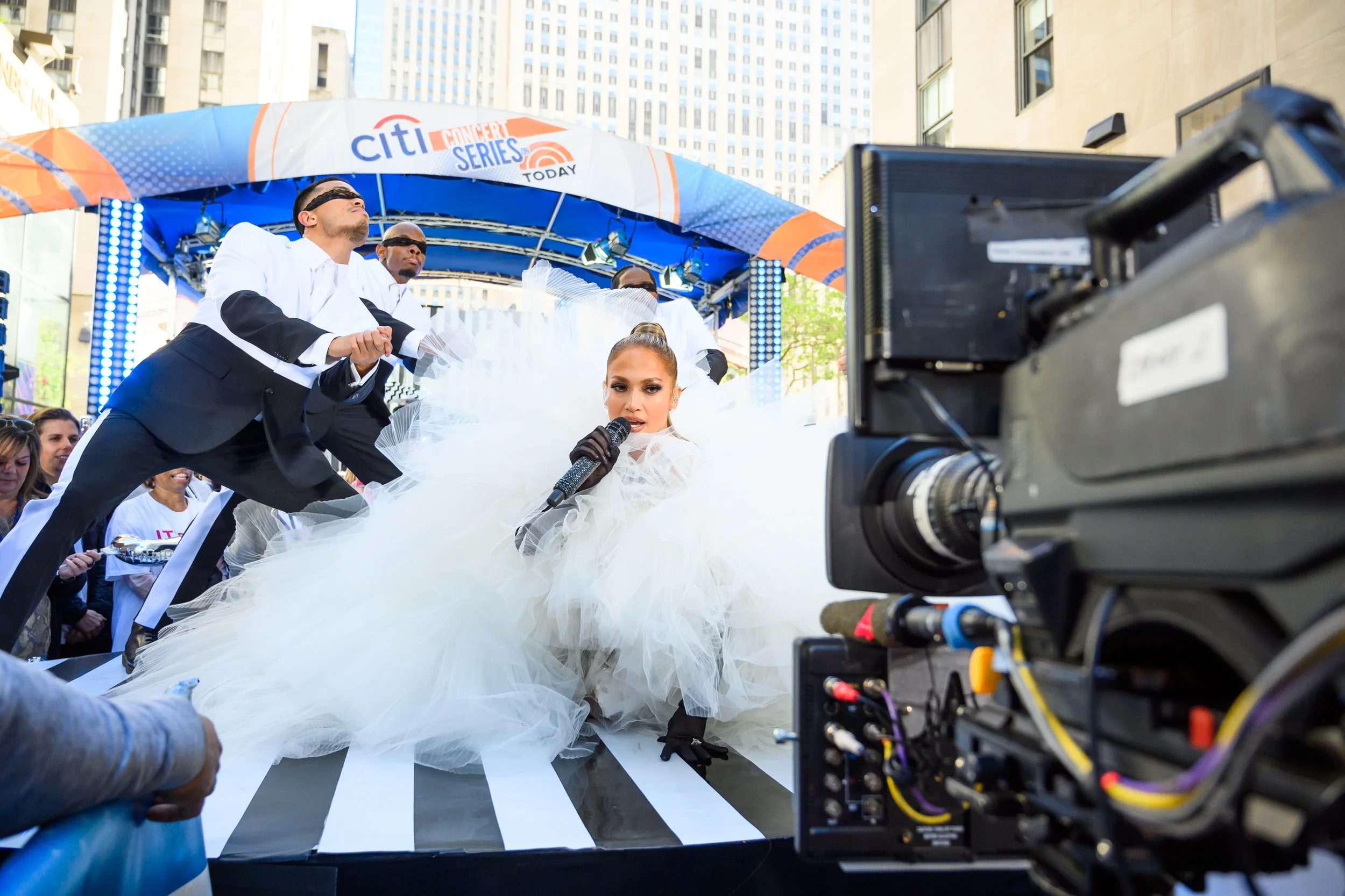 Performers on a street stage during a Citi concert series feature a woman in a white, dramatic gown singing into a microphone, surrounded by dancers in formal black and white tuxedos, with a camera and audience in the background.