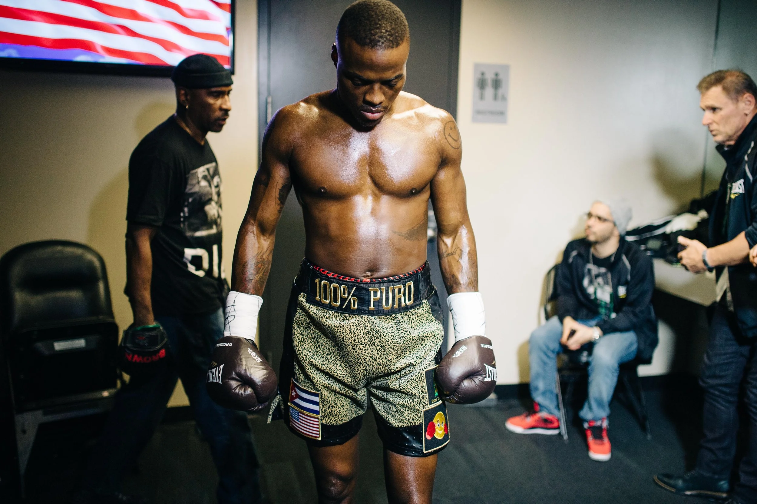 A shirtless male boxer with gold and black shorts, white hand wraps, and boxing gloves, standing with head bowed in a locker room with others around him.