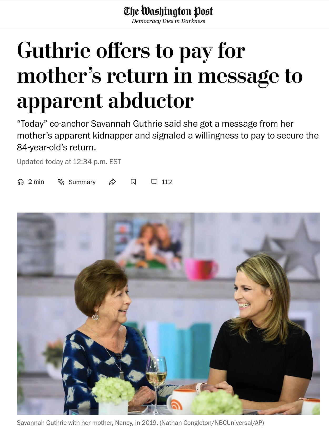 Savannah Guthrie with her mother, Nancy, smiling and talking during a television appearance. They are seated at a table with green floral arrangements, a glass of white wine, and orange and white mugs. The background includes a blurred photo of two c