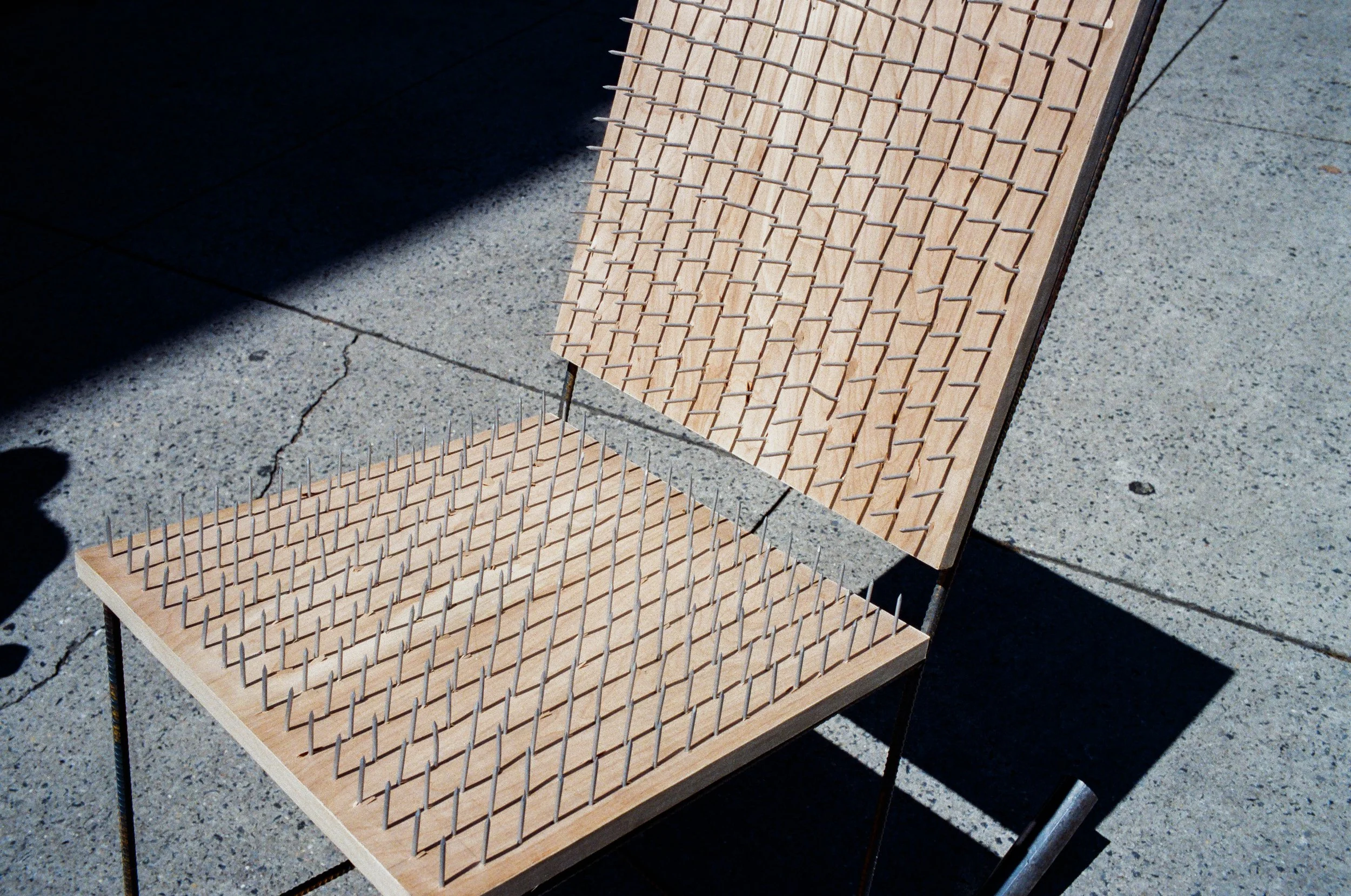 A wooden chair with a backrest and seat filled with numerous metal spikes, set outdoors on a concrete sidewalk, casting a shadow.