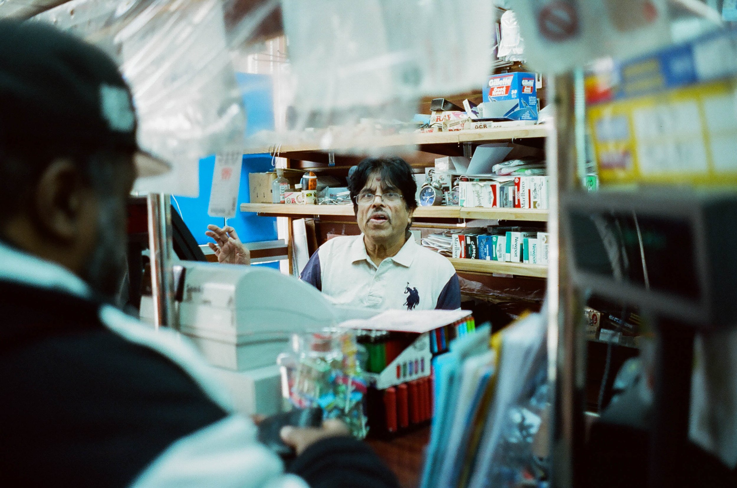 Interior of a small store with shelves stocked with various products. Two men are engaged in conversation; one behind the counter, another in front, visible from behind. The man behind the counter has black hair and glasses, wearing a black and white