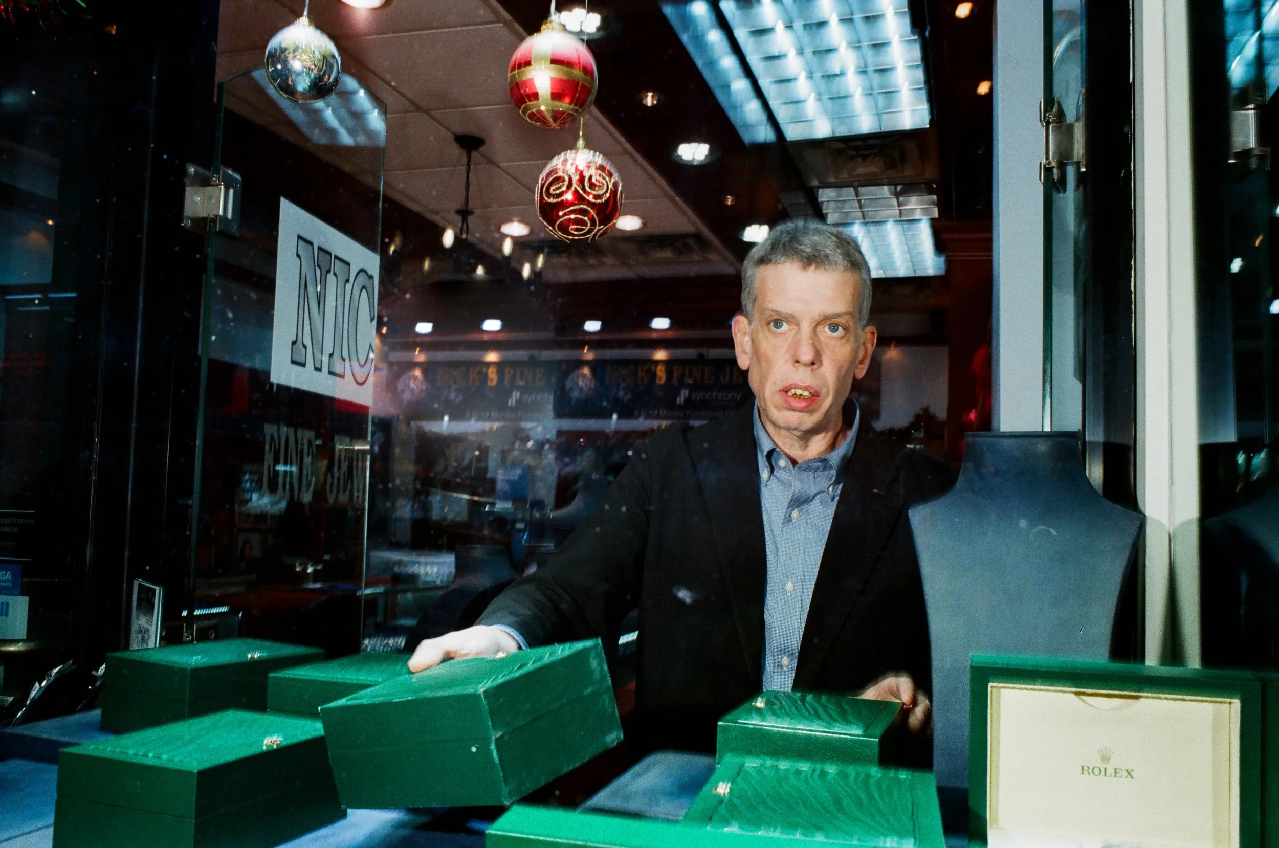 Man with short hair and light skin in a dark blazer and blue shirt inside a store, handling multiple green boxes with a Rolex box in front, decorated with Christmas ornaments hanging from the ceiling
