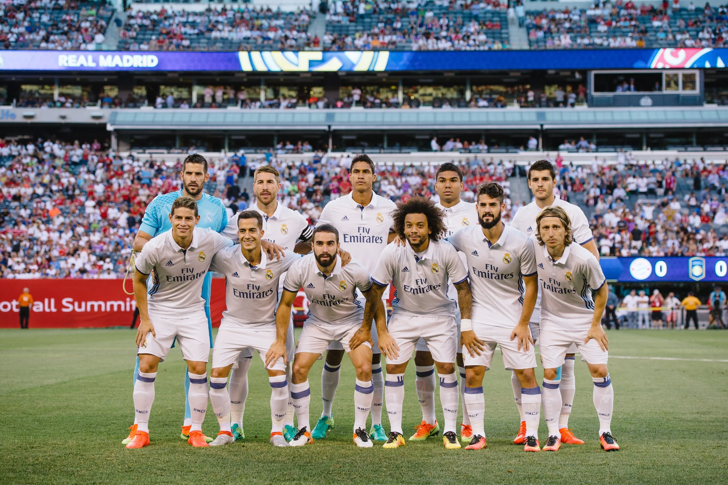A soccer team in white uniforms with 'Fly Emirates' logo posing for a team photo on the field before a match at a stadium filled with spectators.