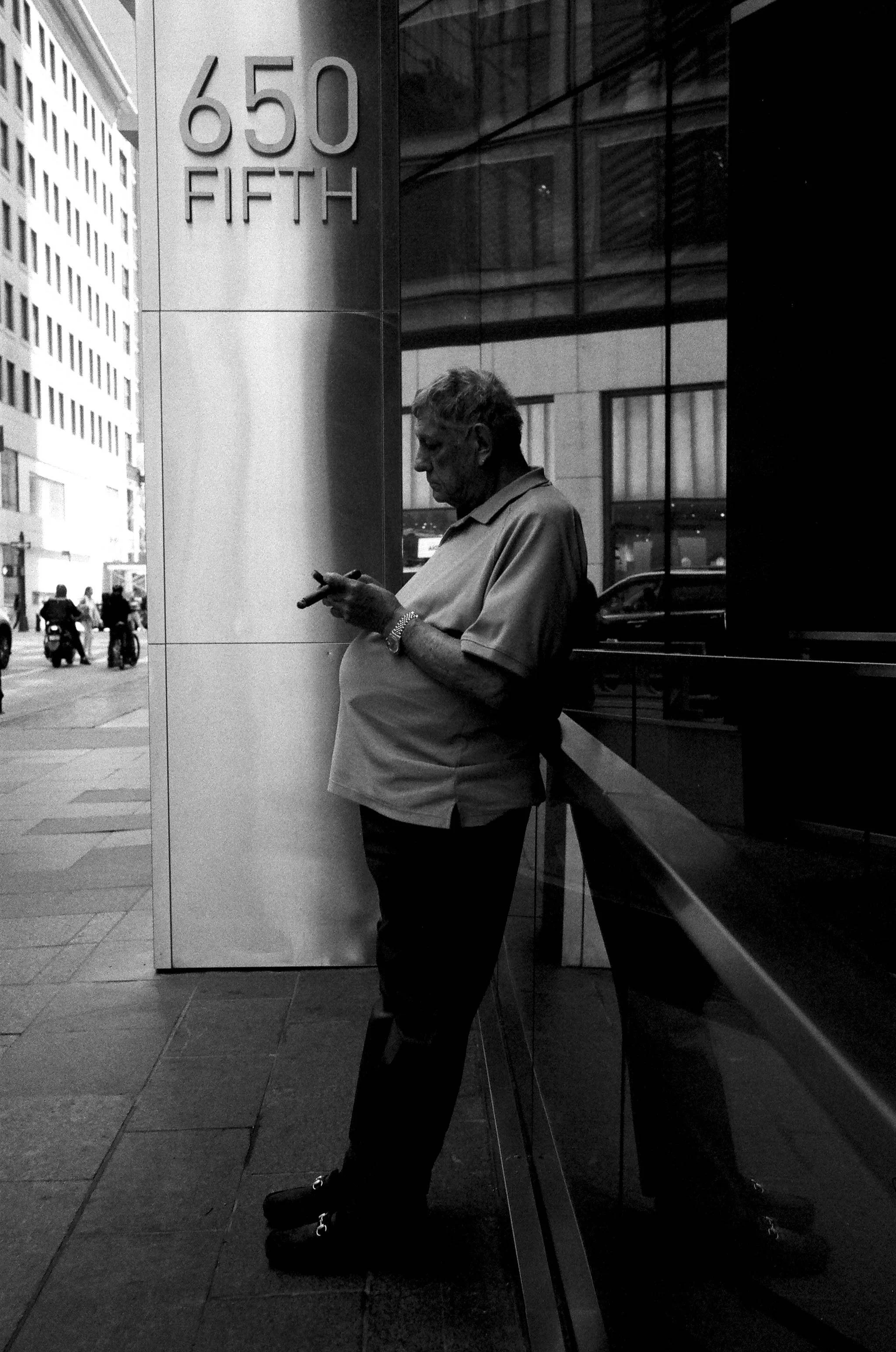 A man leaning against a reflective glass building while looking at his phone on a city sidewalk, with street and traffic visible in the background, 650 Fifth Avenue.