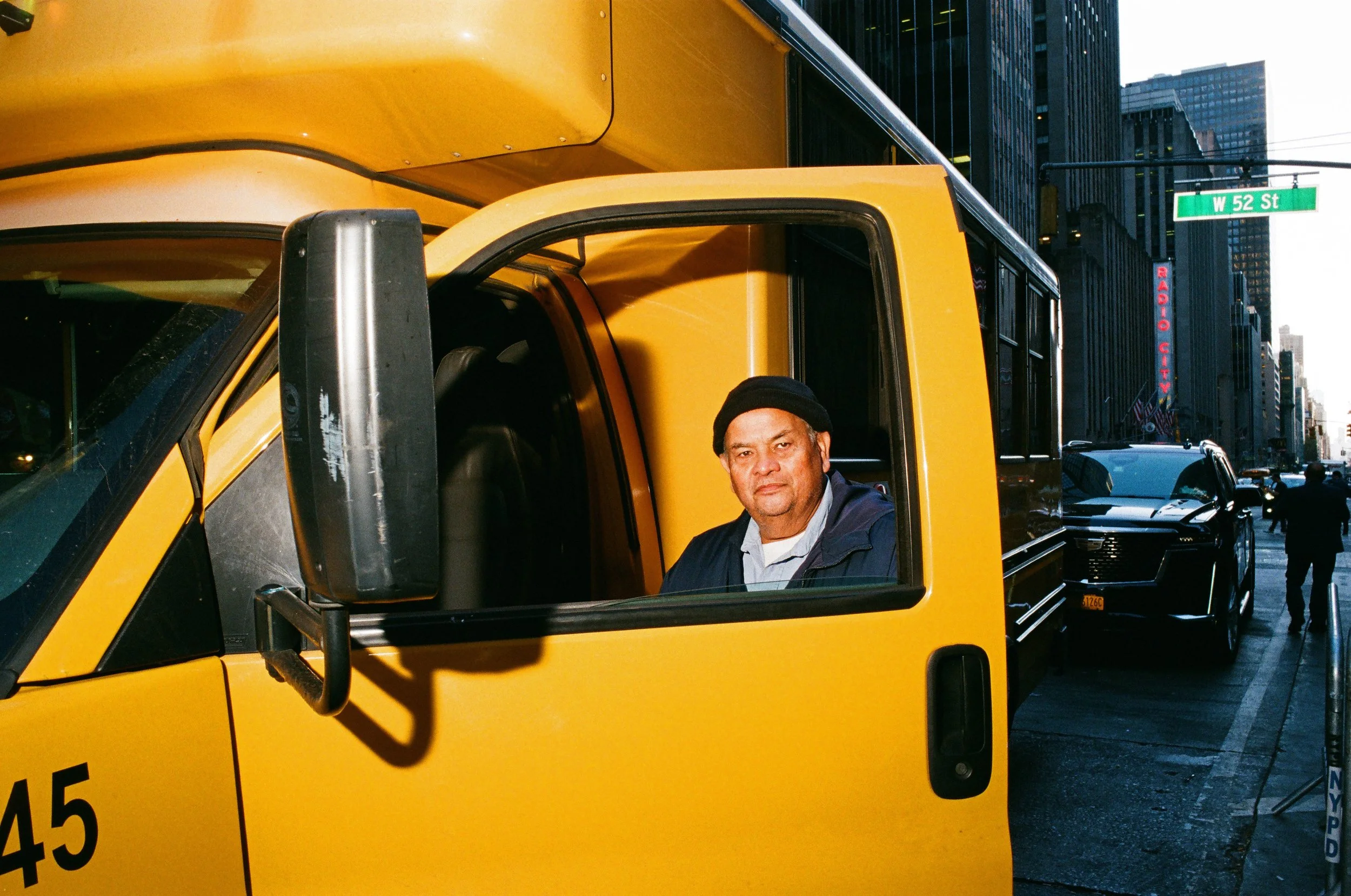 A man sitting in the driver's seat of a yellow delivery truck on a city street with tall buildings and a street sign that reads W 52 St.