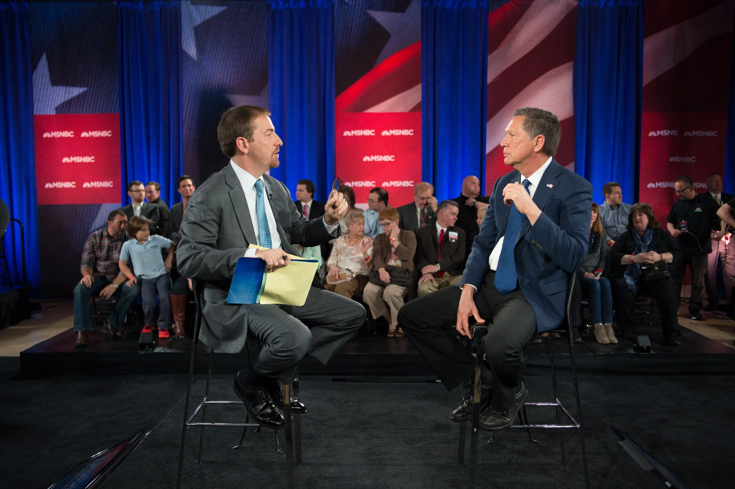 Two men in suits having a discussion on a stage during a televised political debate, with a background of blue curtains and an audience seated behind and around them, and MSNBC banners on the backdrop.