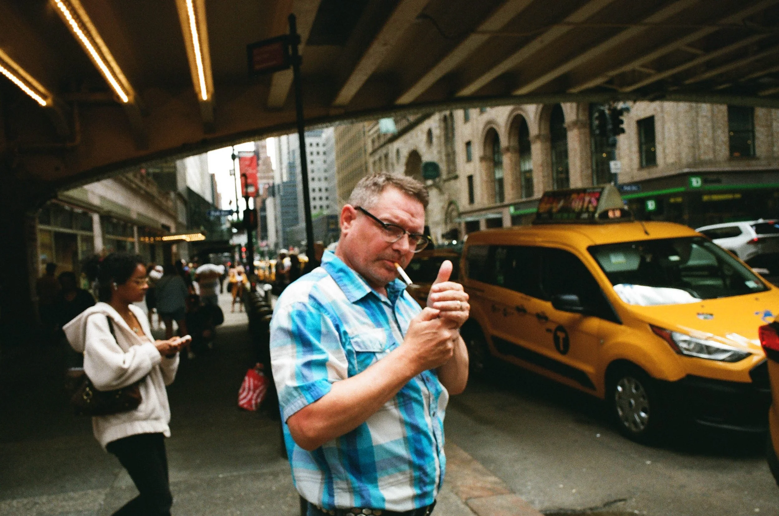 A man with glasses and a blue plaid shirt lighting a cigarette on a busy city street with yellow taxis and pedestrians under an overpass.