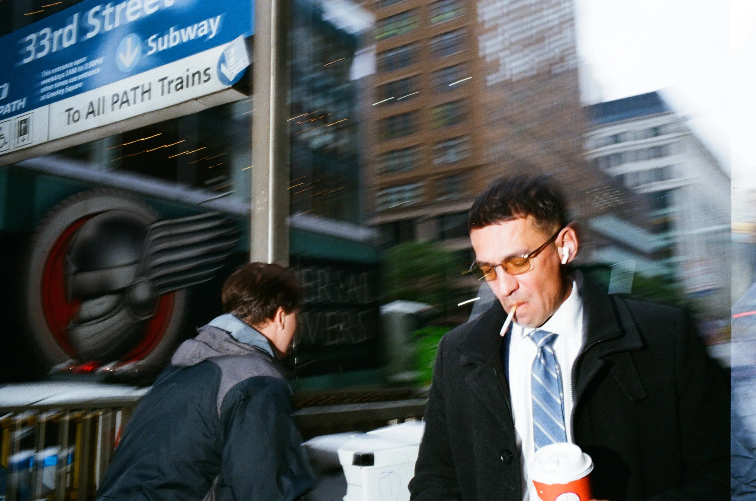 A man with sunglasses, a cigarette, and a coat holding a coffee cup walking on a city sidewalk, with buildings and a street sign for 33rd Street Subway in the background.
