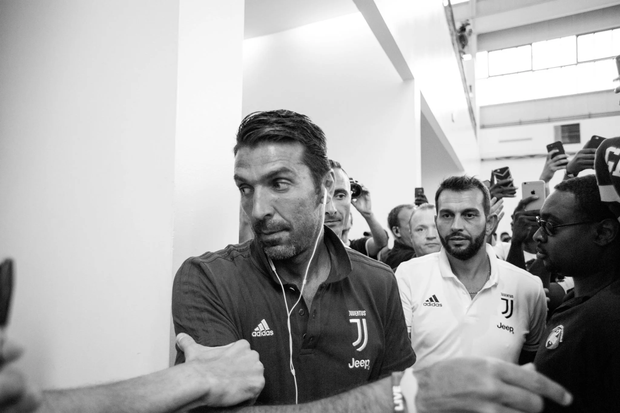 Black and white photo of a man in a Juventus Juventus football team shirt, surrounded by people, with some taking photos or videos, indoors near a white wall and ceiling.