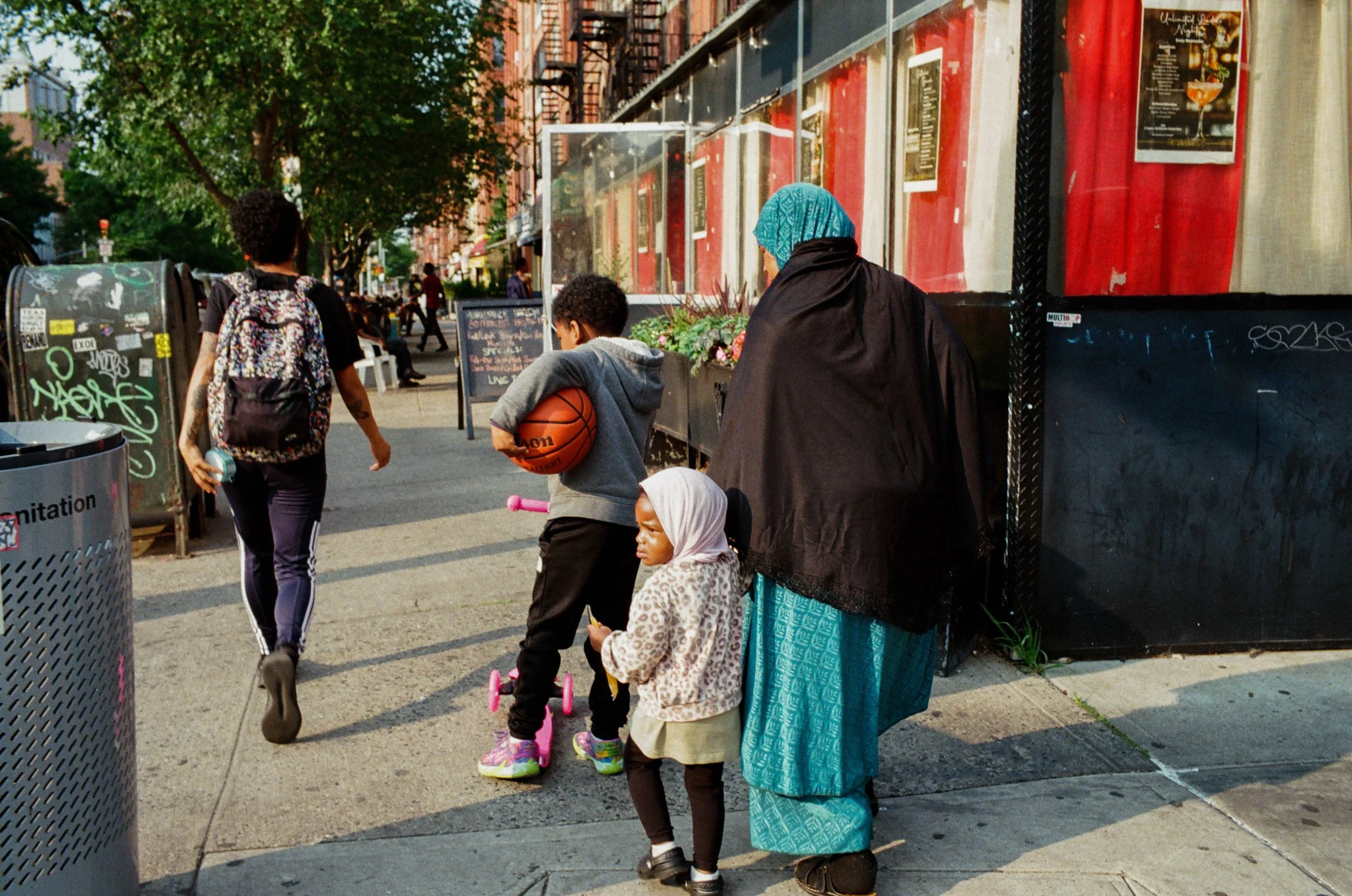 People walking on an urban sidewalk in front of a restaurant. A woman with a blue headscarf and a long teal skirt stands with two children, one holding a basketball and the other on a pink skateboard. A young person with a backpack walks ahead, carry