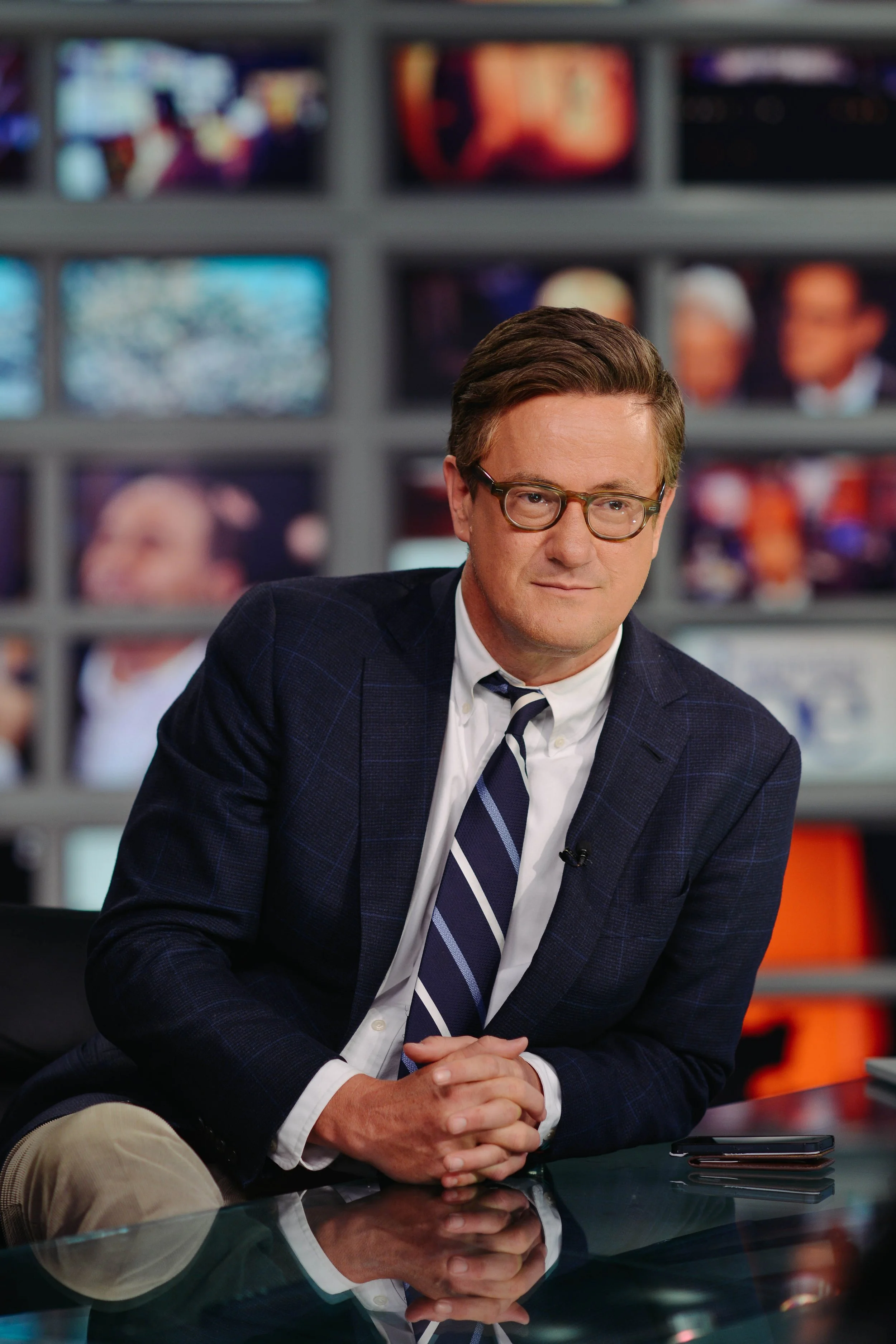 A man in a navy checkered blazer, white dress shirt, and striped tie, sitting at a newsdesk with multiple TV screens in the background.