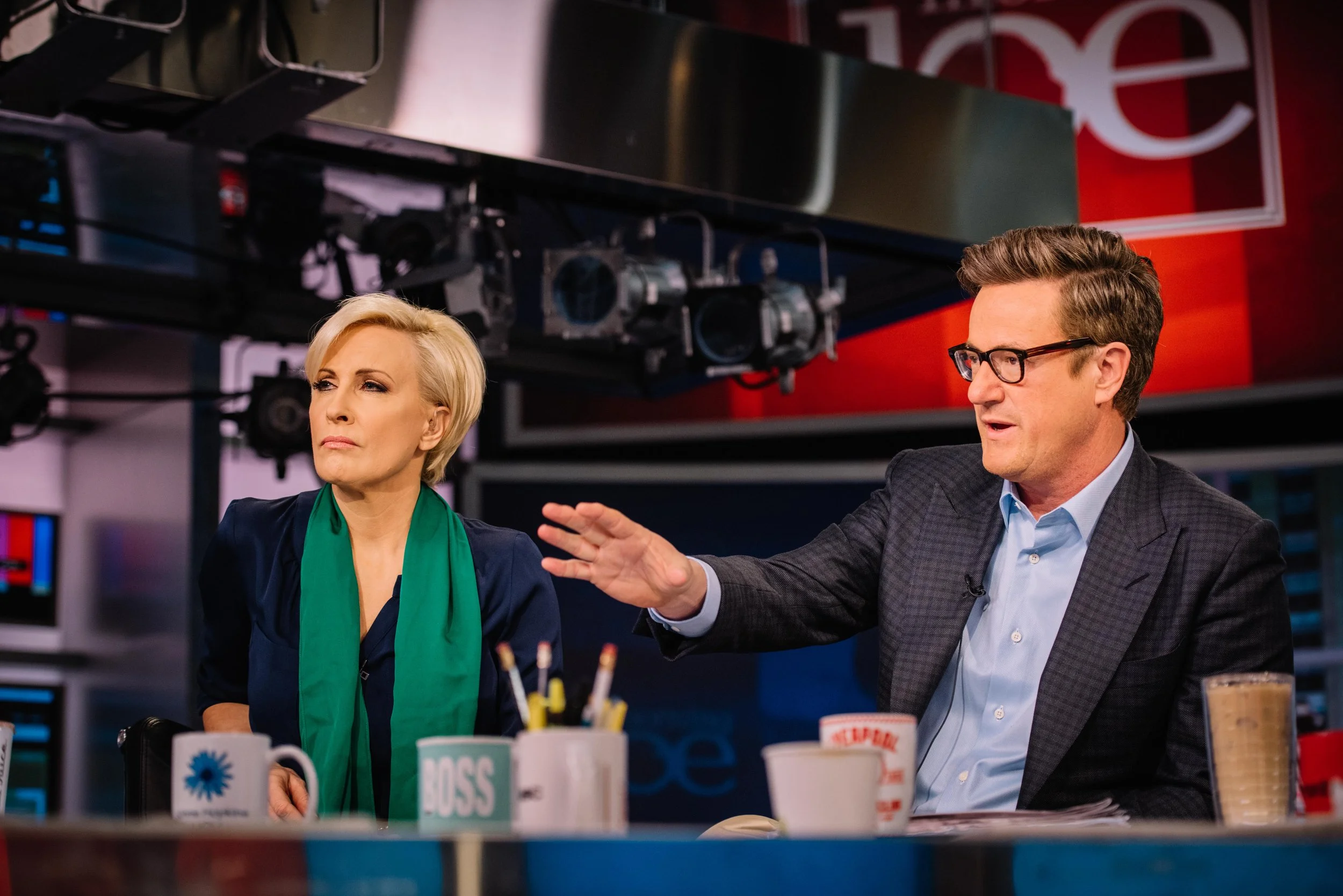 Two news anchors sit at a table in a news studio. The woman has short blonde hair and wears a navy blouse with a green scarf. The man has brown hair, glasses, and wears a checked blazer over a light blue shirt. There are mugs, a cup of coffee, and a 
