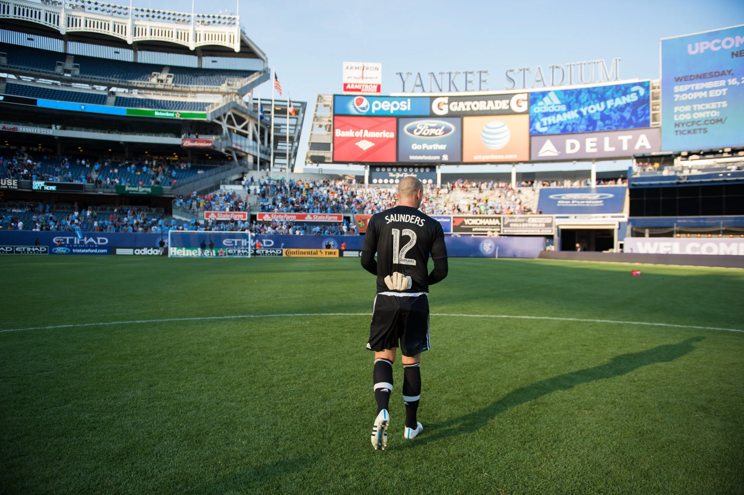 A soccer player standing on the field with his back to the camera at Yankee Stadium, with the crowd and electronic billboards in the background.