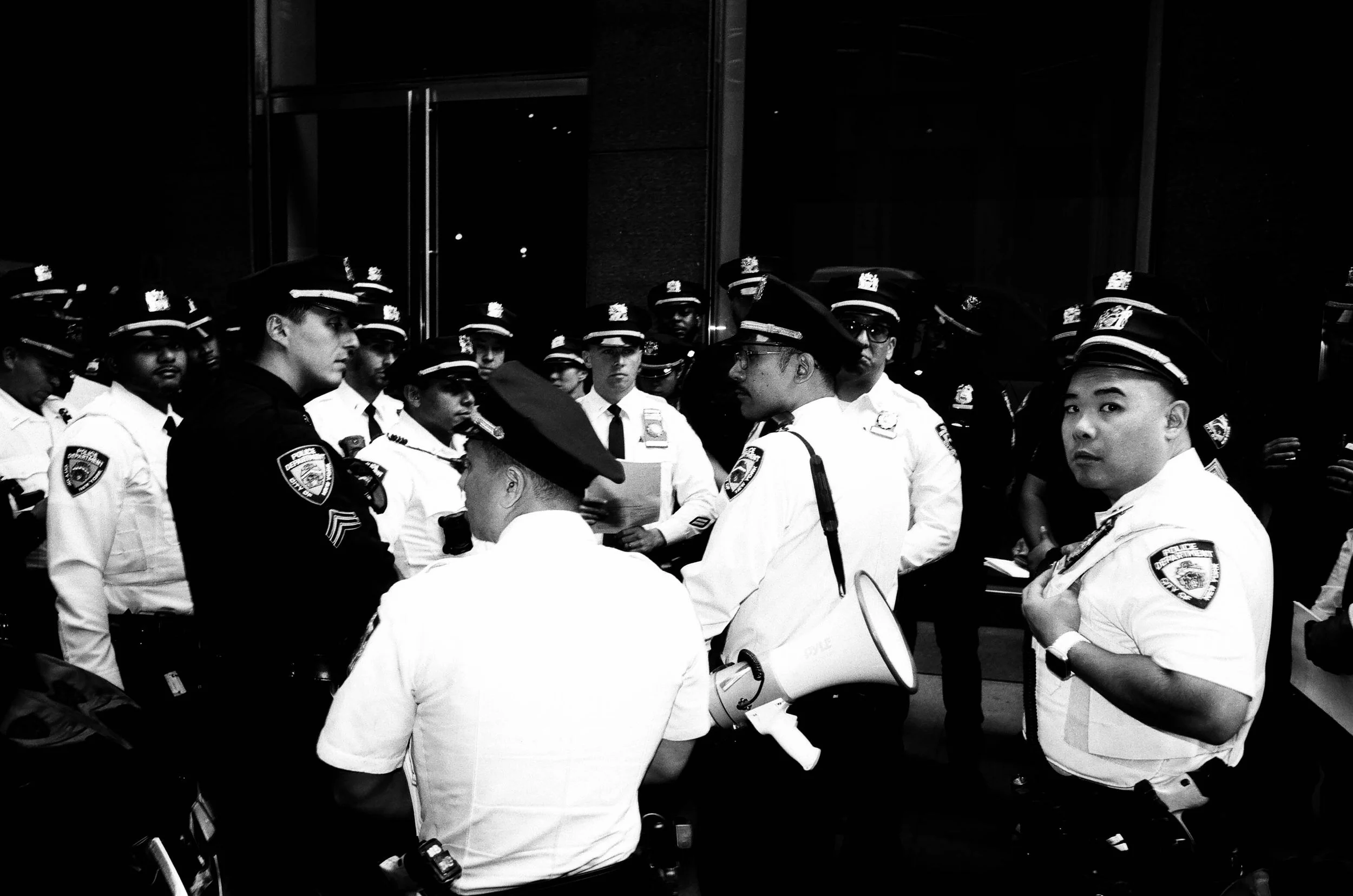 A group of police officers gathered in a circle, some holding papers and a megaphone, with a reflection of lights on the glass behind them.