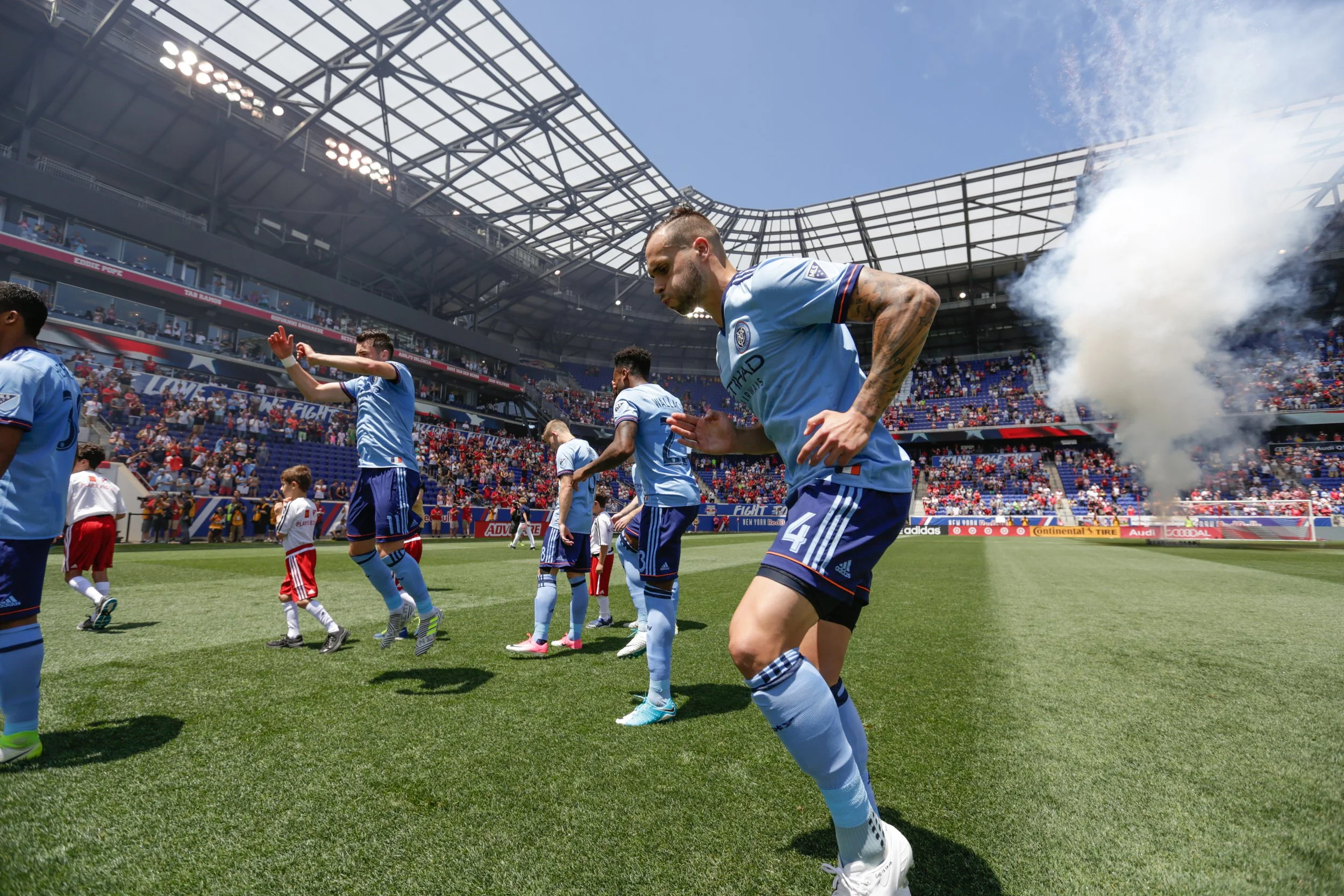 Soccer players in light blue uniforms entering the field for a match in a large stadium filled with fans.