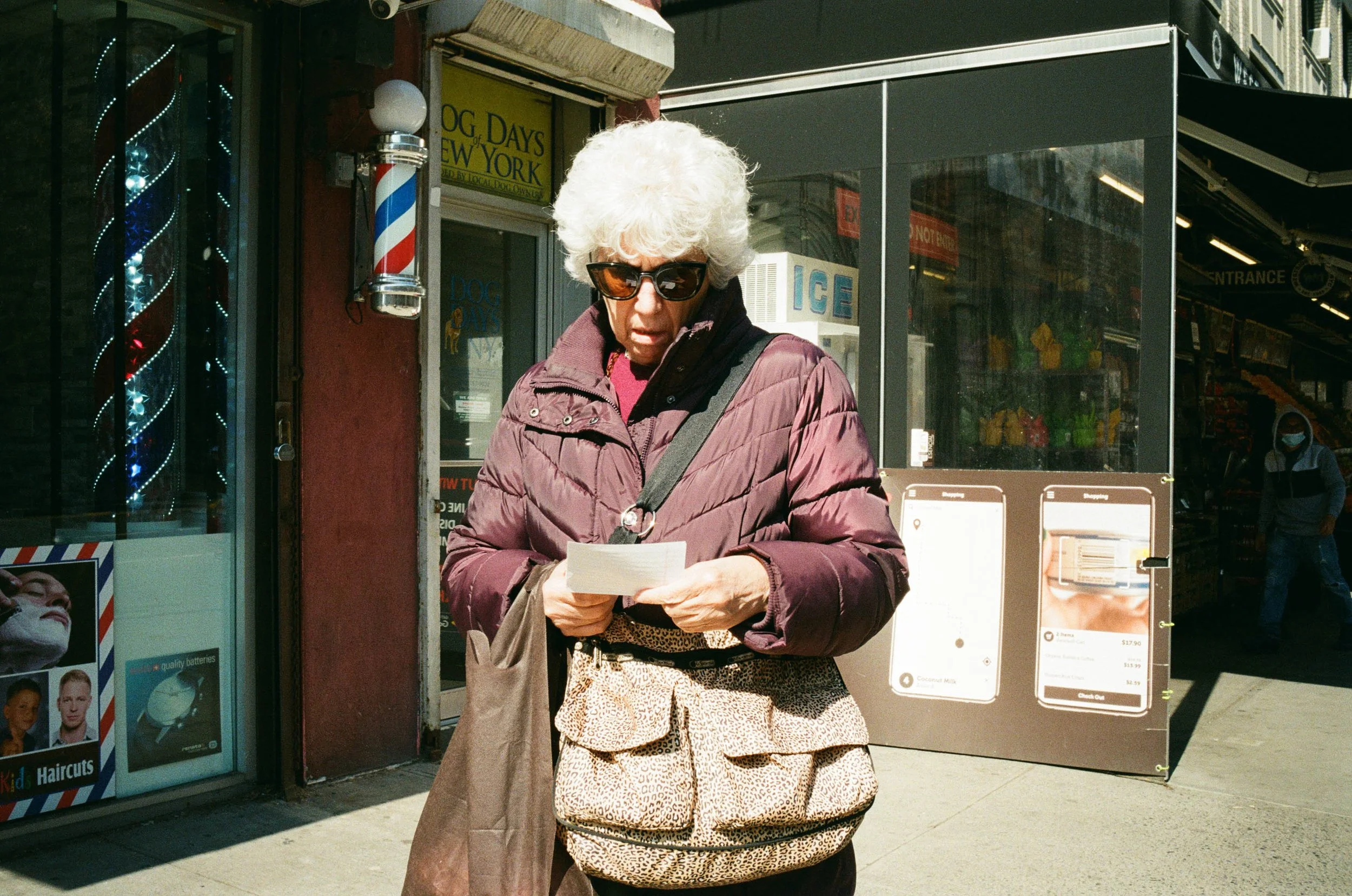 An elderly woman with white curly hair wearing sunglasses, a purple jacket, and a leopard print bag, looking at a piece of paper on a city sidewalk in front of a storefront with a barber pole and behind a glass-enclosed ice machine, with a person wea
