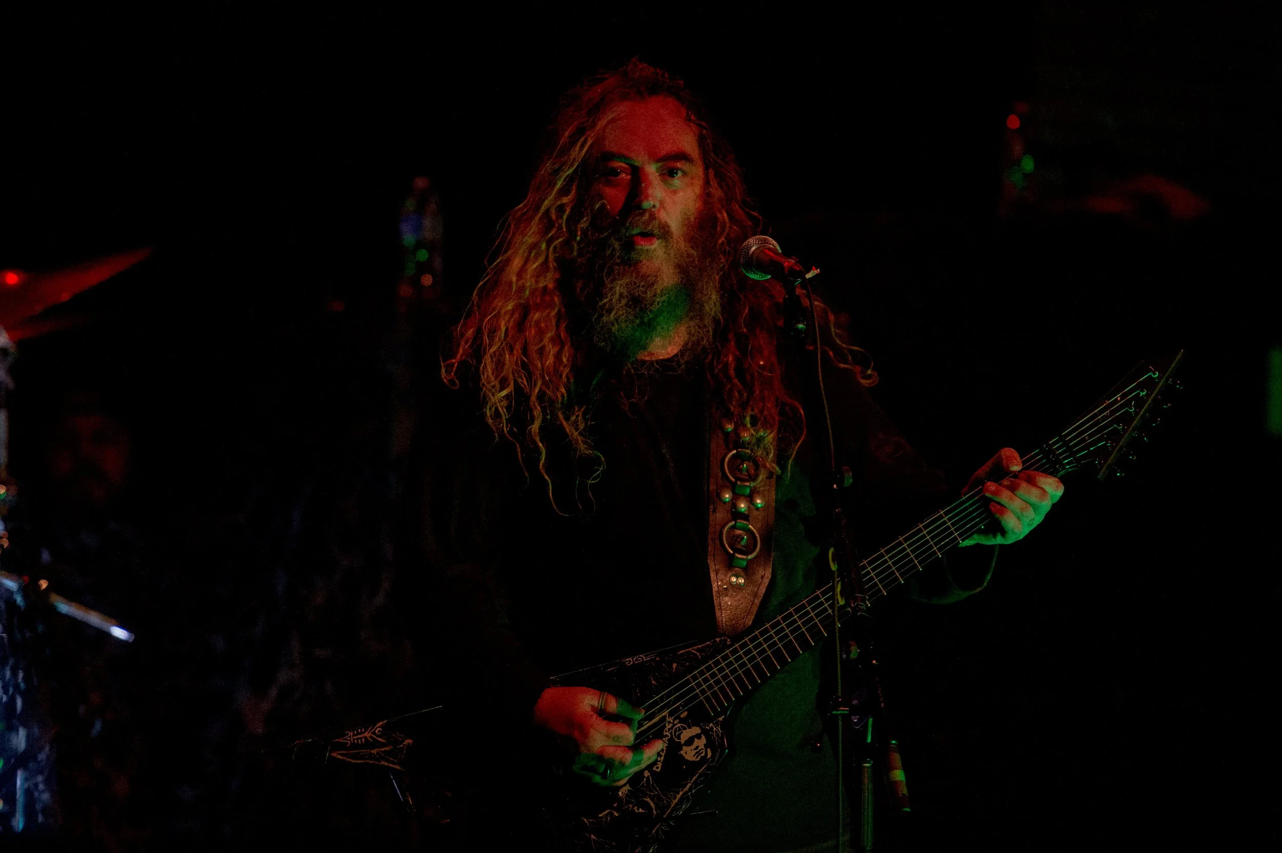 Musician with long curly hair and beard playing electric guitar on stage with microphone, dark background, dim colorful lighting.