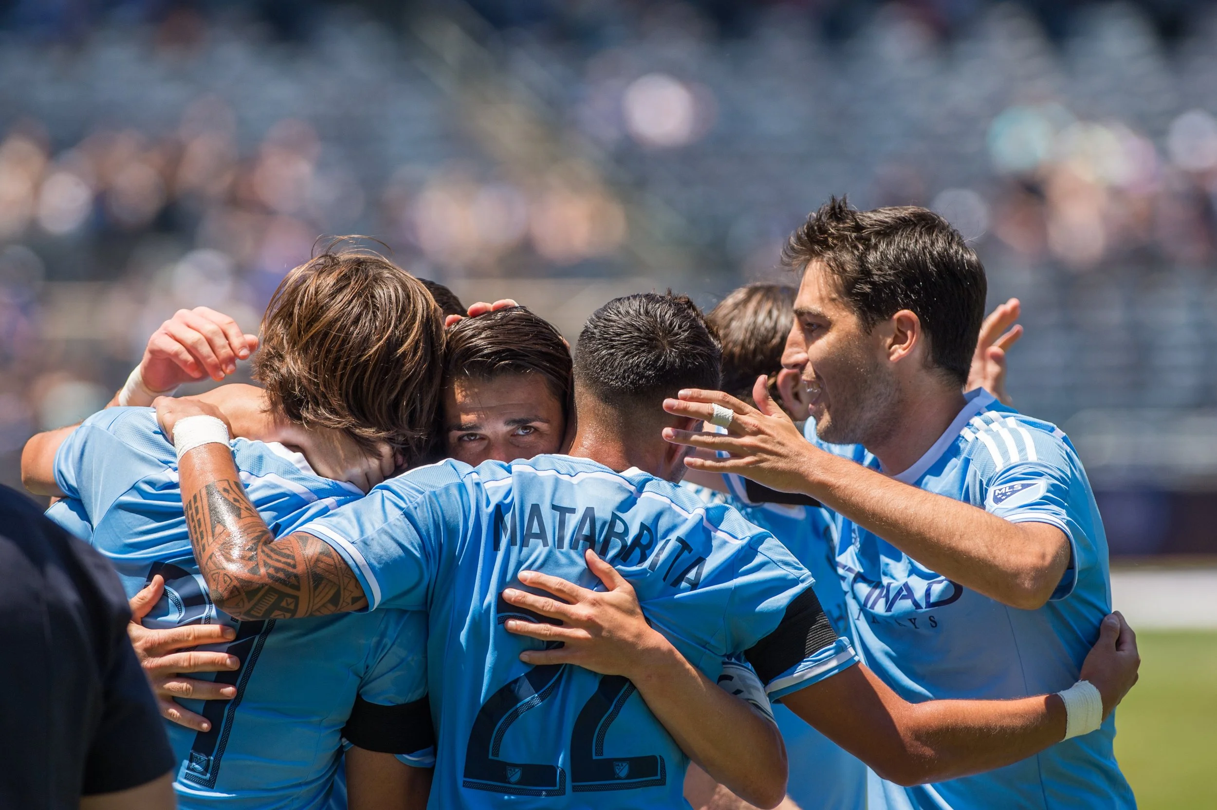 Soccer players in light blue jerseys celebrating a goal by hugging each other on the field.
