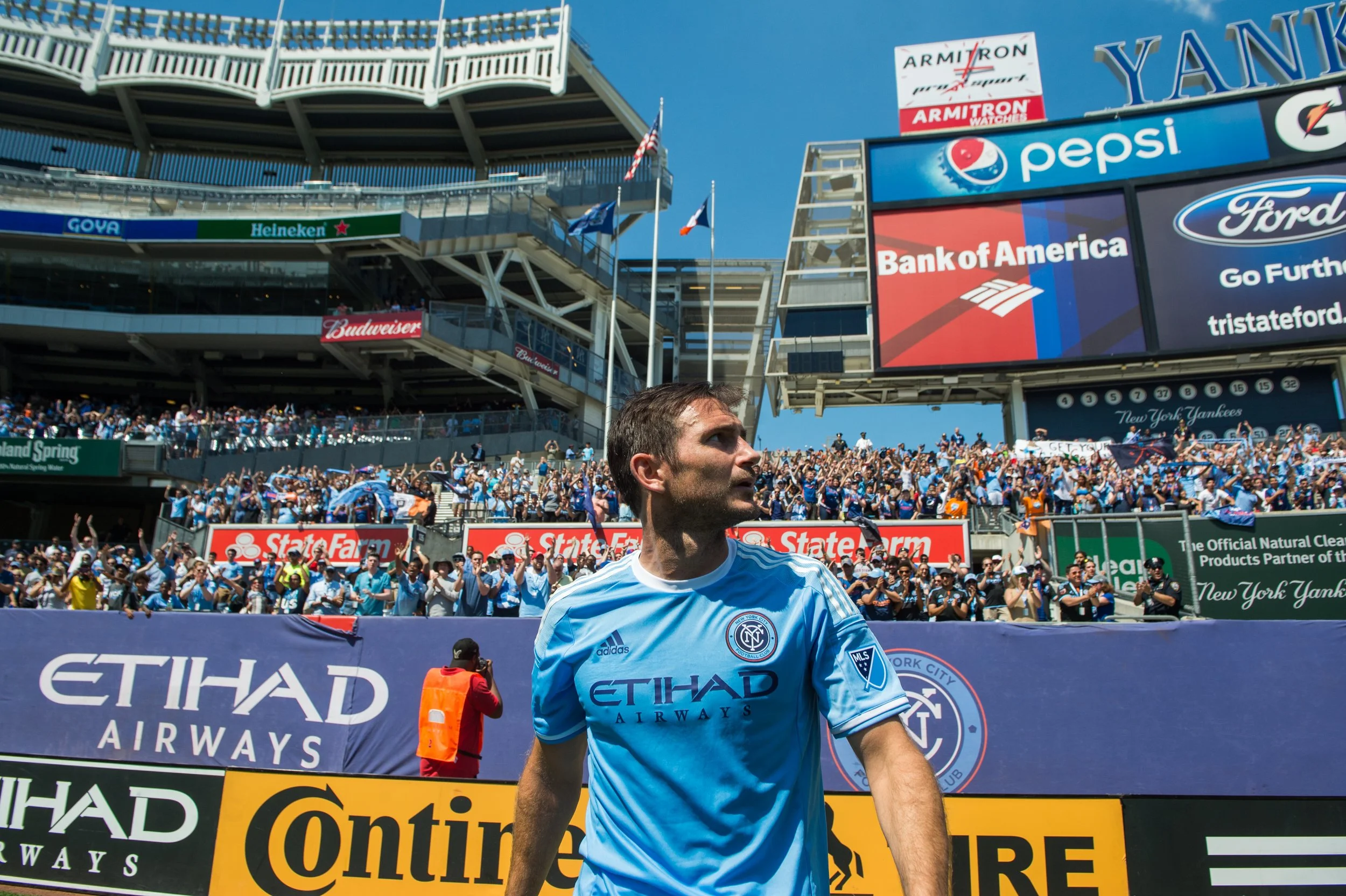 A soccer player wearing a light blue New York City FC jersey with the Etihad Airways logo, walking on the field in front of a large crowd at Yankee Stadium, with scoreboard and advertisements in the background.