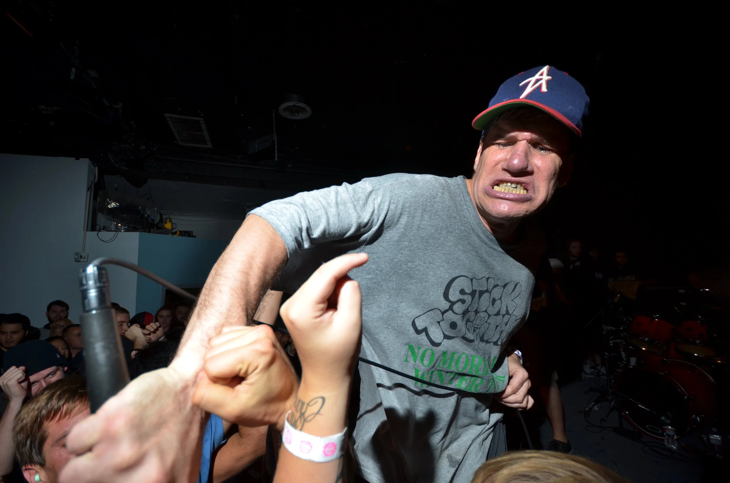 A man with on a navy baseball cap with a red brim, wearing a gray T-shirt, showing a tense facial expression during a concert or event, surrounded by a crowd of people.