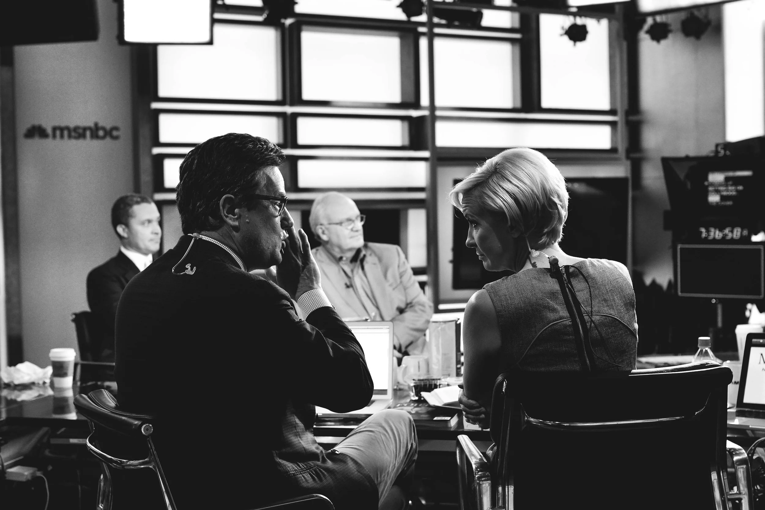 Black and white photo of a television news studio with four people engaged in a conversation or interview. There are multiple monitors and equipment around them. One woman with short blonde hair is facing a man with glasses and dark hair, who is list