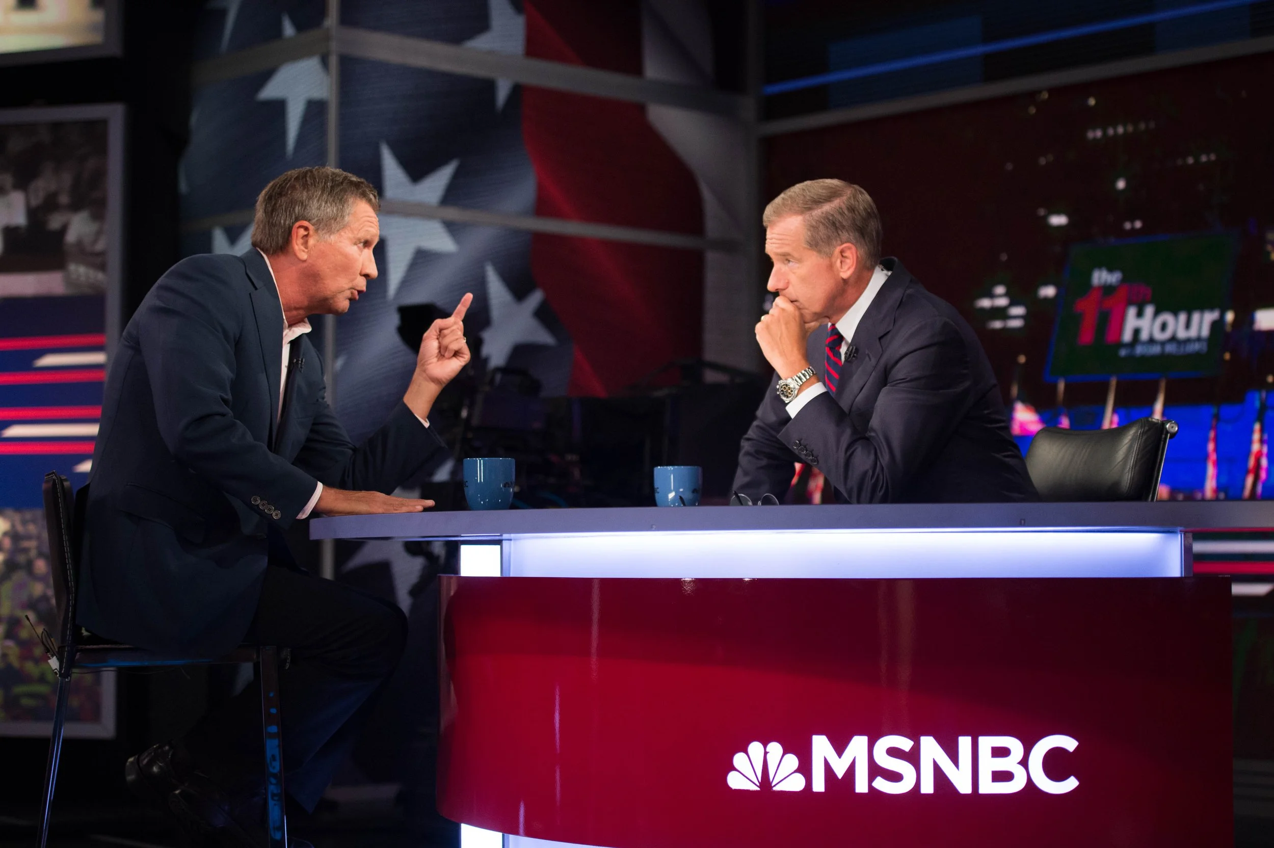Two men having a discussion during a TV debate on MSNBC studio, with American flag and political graphics in background.