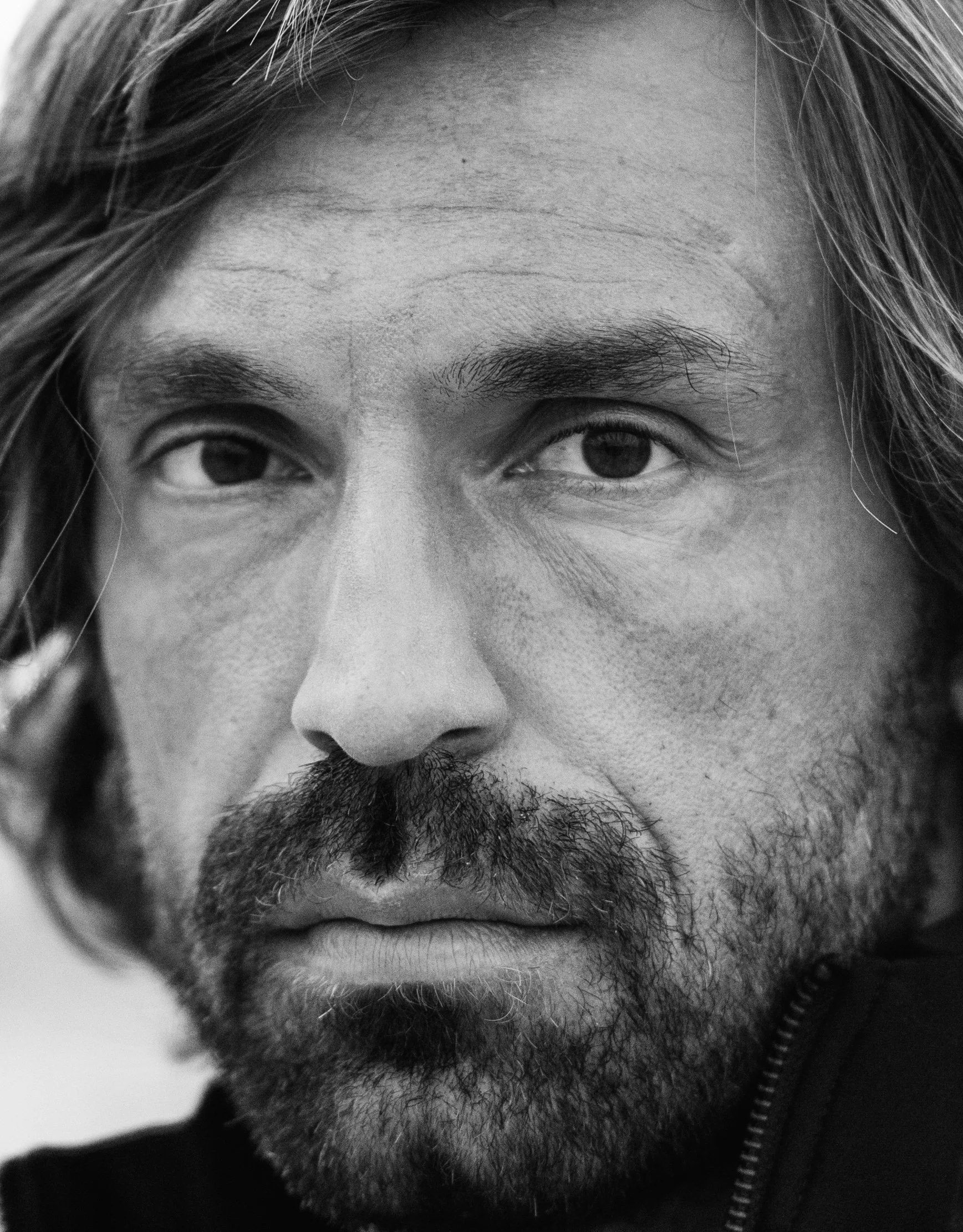 Close-up black and white photo of a man's face with long hair and beard, staring directly into the camera.