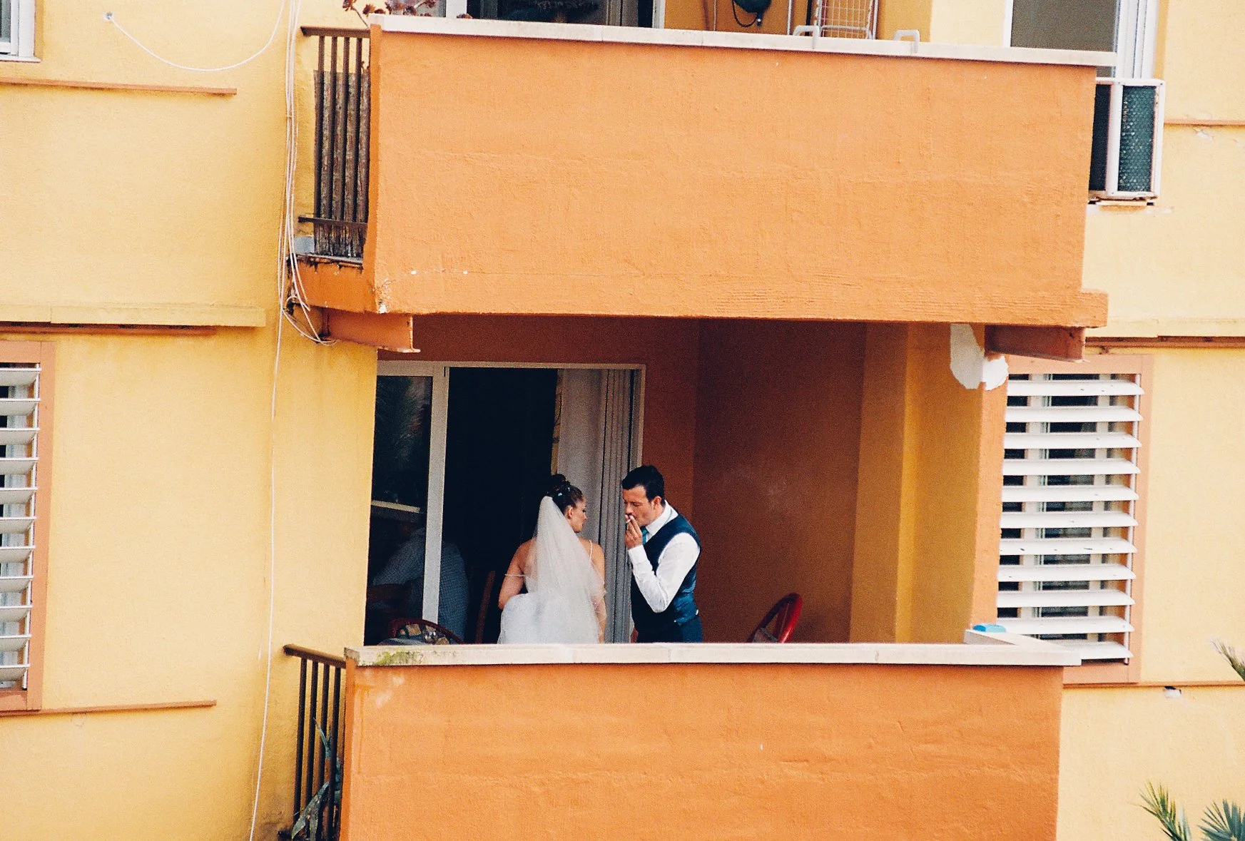 A bride and groom on a balcony, with the groom whispering to the bride, inside an orange and yellow building.