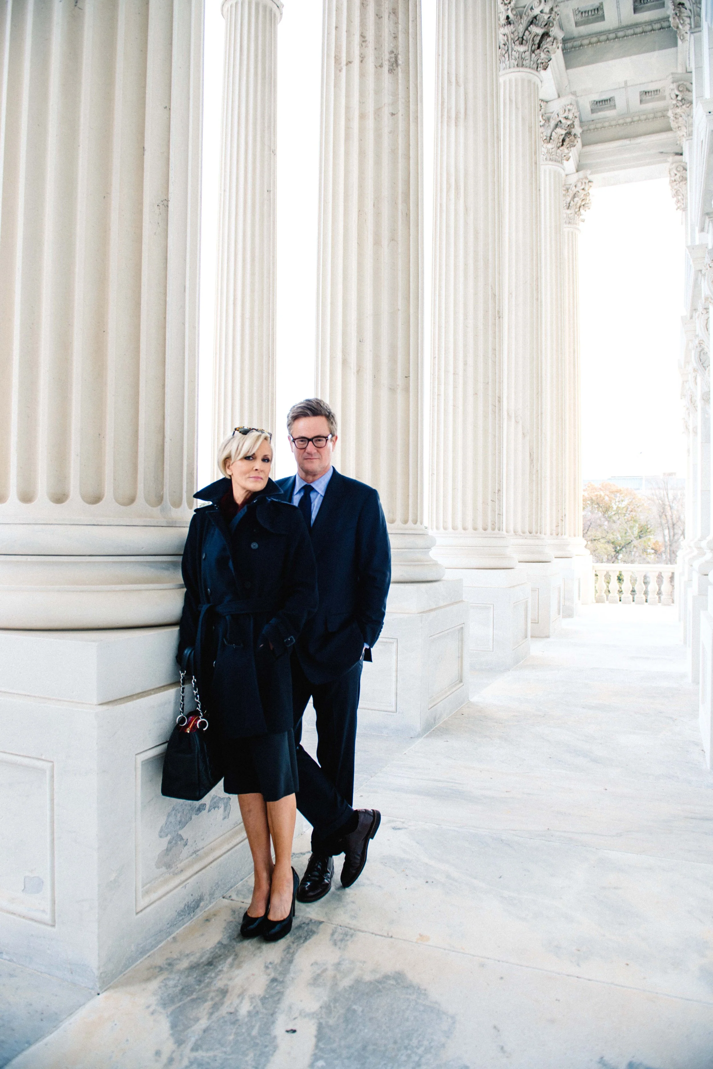 A man and woman dressed in formal black attire standing against large white classical columns on a marble building porch.