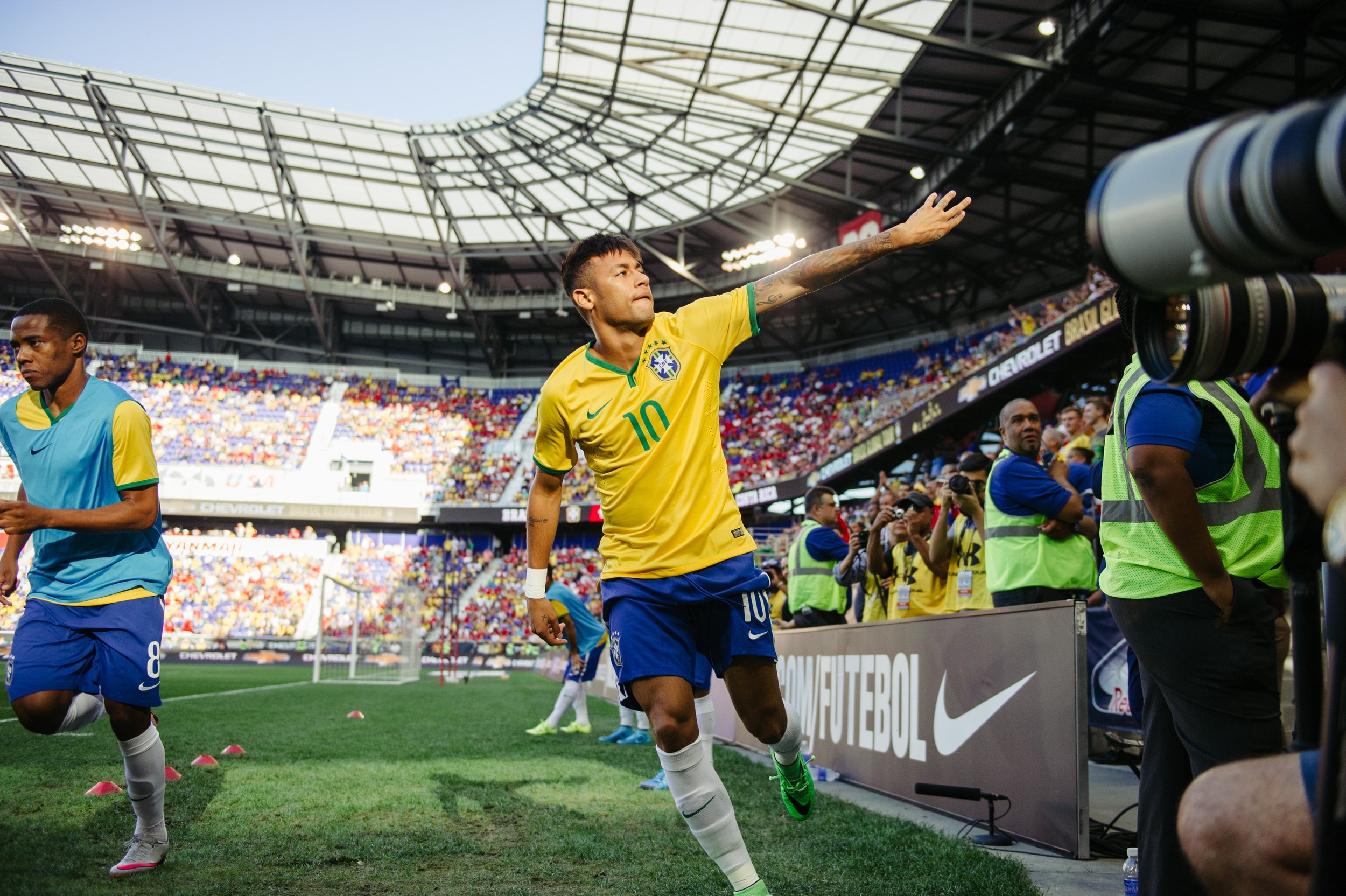 Brazilian soccer player celebrating on the field wearing a yellow jersey with number 10, surrounded by teammates and photographers in a stadium filled with spectators.