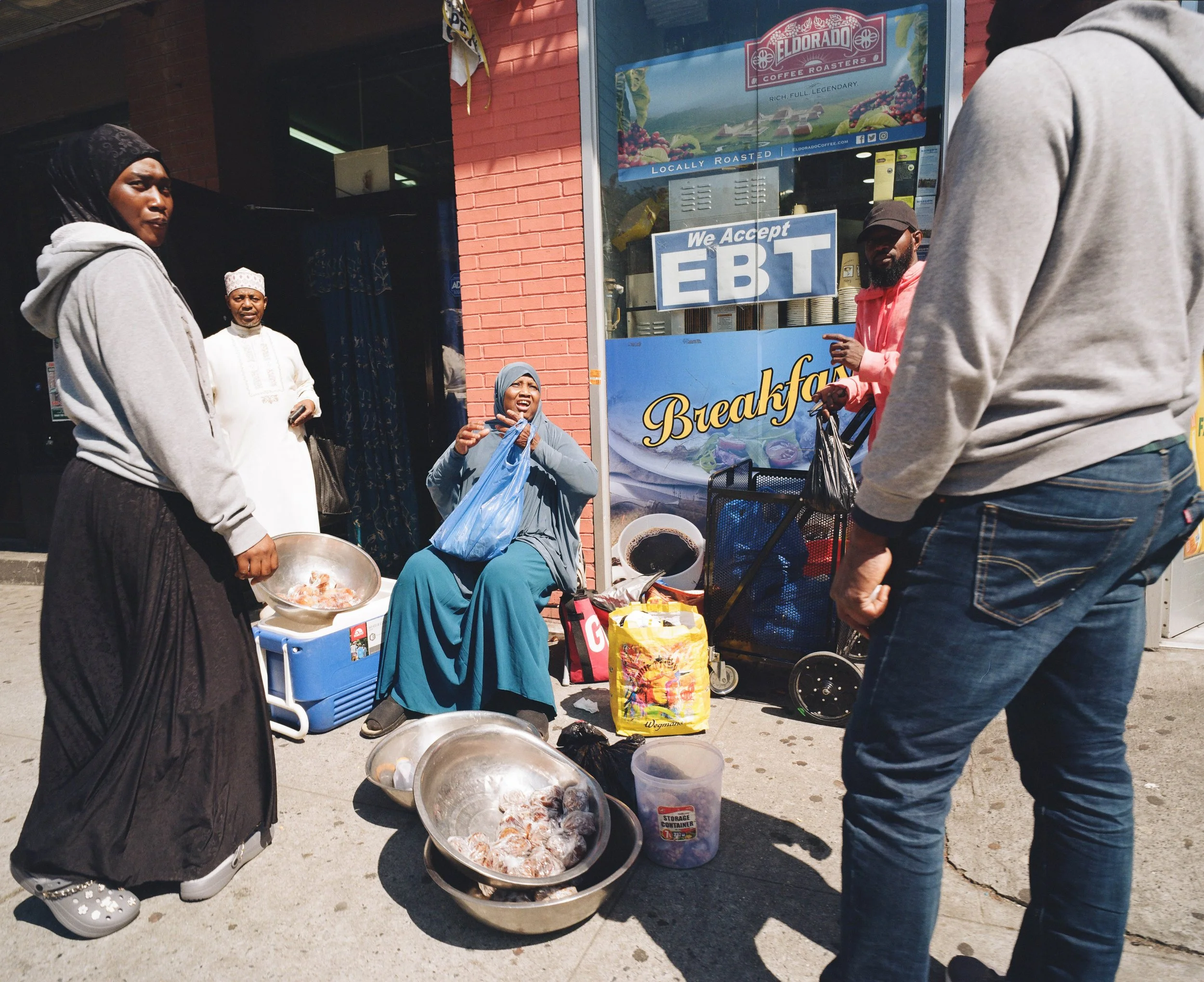 A group of people gathered outside a coffee shop, some selling seafood, with large bowls of seafood on the ground. A woman sitting on a chair is smiling and holding a blue plastic bag, while others stand nearby. The storefront has signs indicating ac