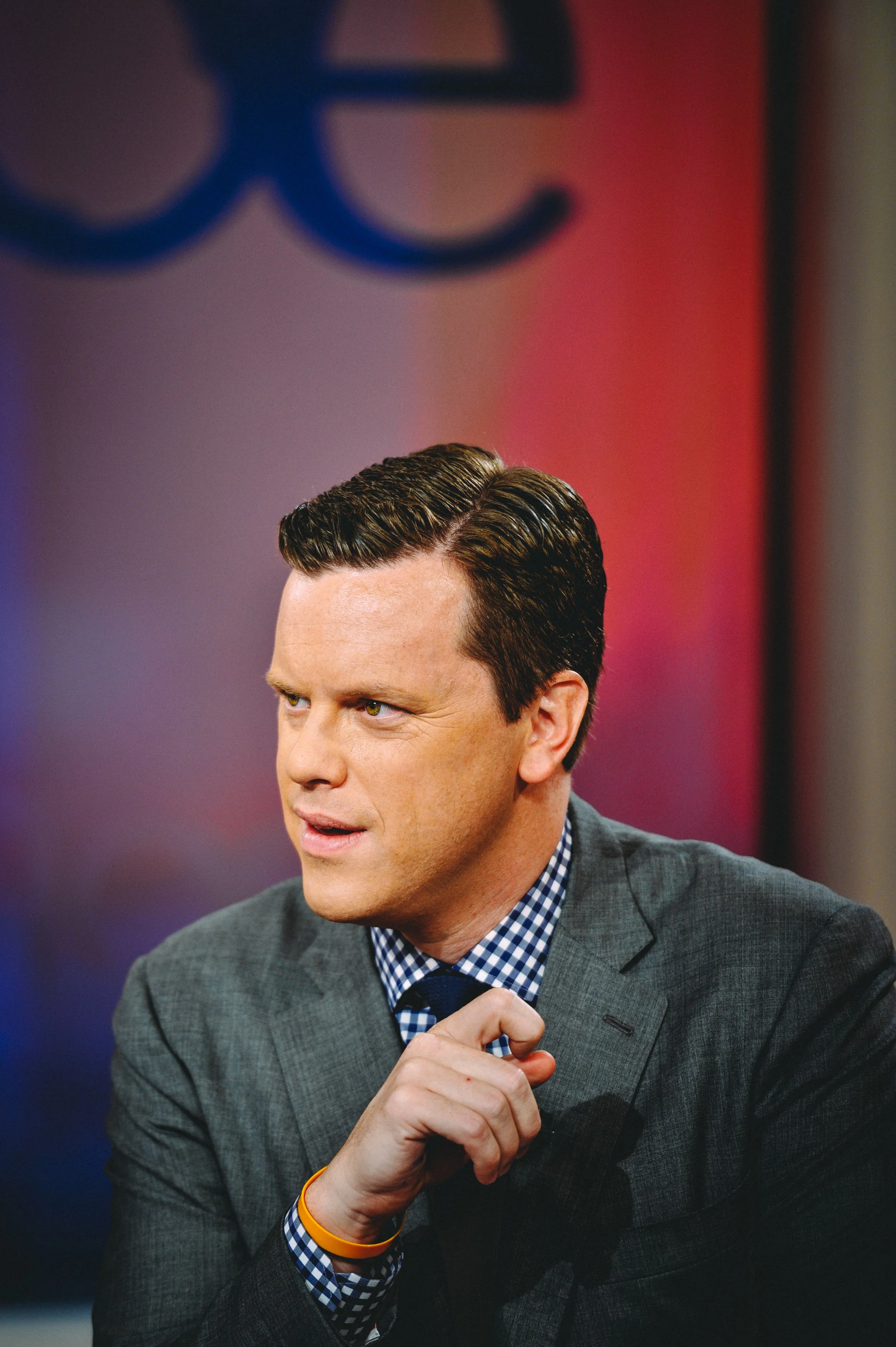 A man in a gray suit with a blue and white checkered shirt gestures with his hand during a TV interview, with a NewsNation logo in the background.