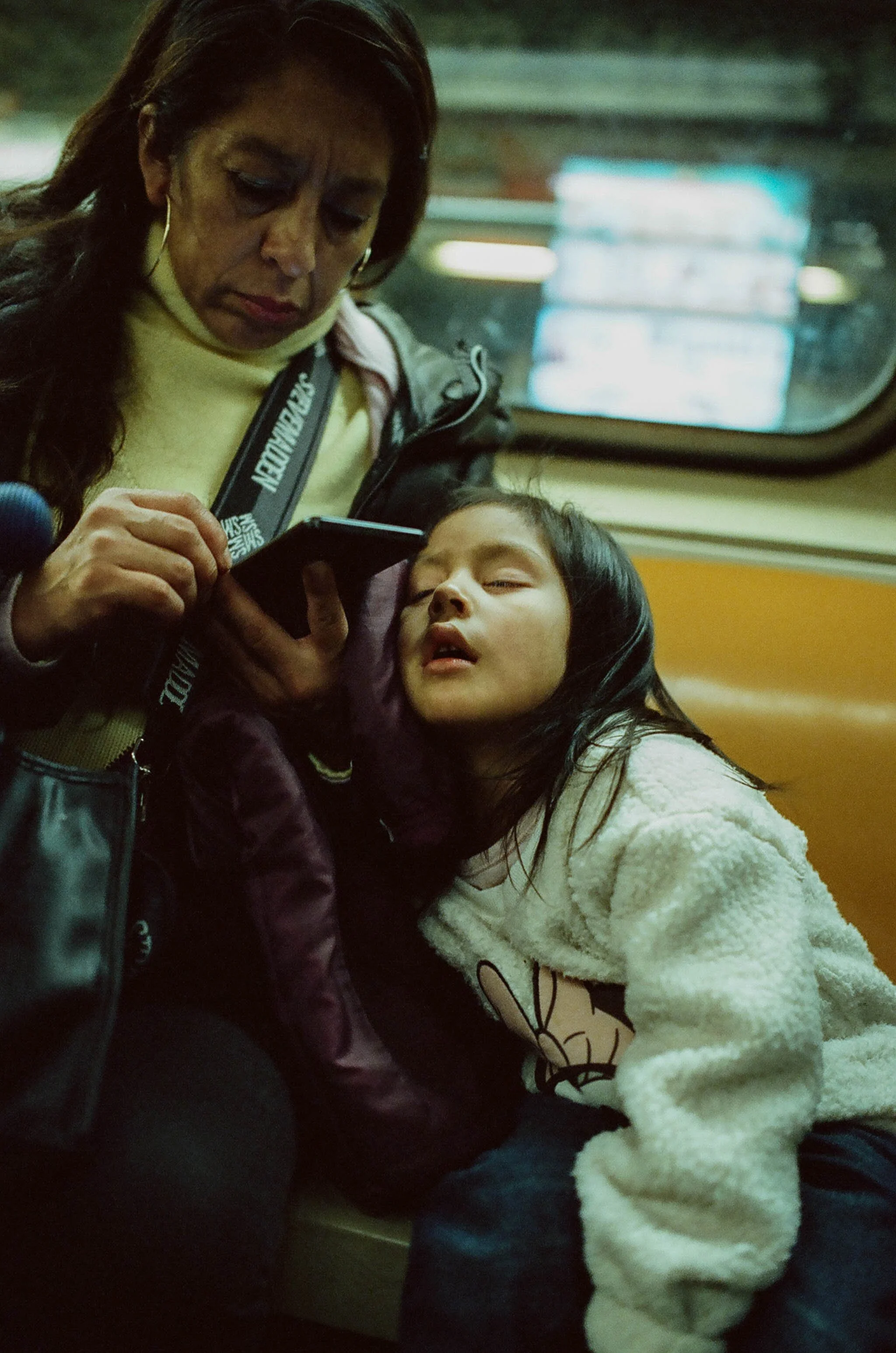 A woman and a young girl are sitting together on a train, with the girl leaning on the woman's chest, both looking at a smartphone.