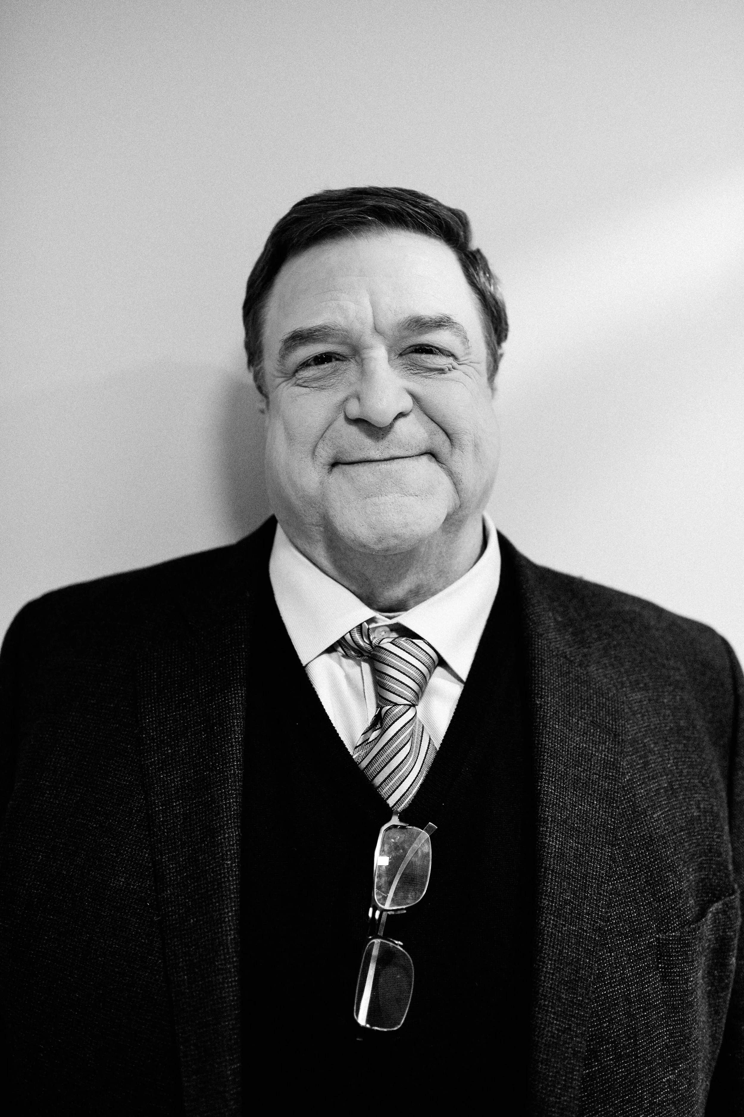 Black and white photo of a smiling middle-aged man with glasses hanging from his collar, wearing a suit and striped tie, standing against a plain wall.