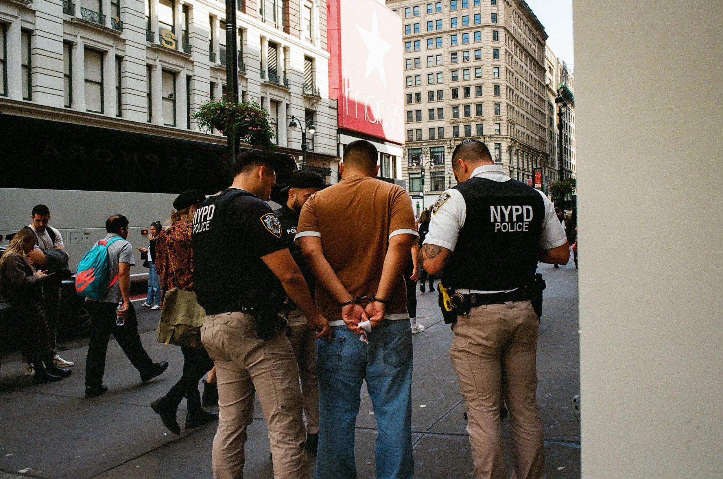 A man in handcuffs is being detained by three NYPD police officers on a city street, with pedestrians walking in the background.
