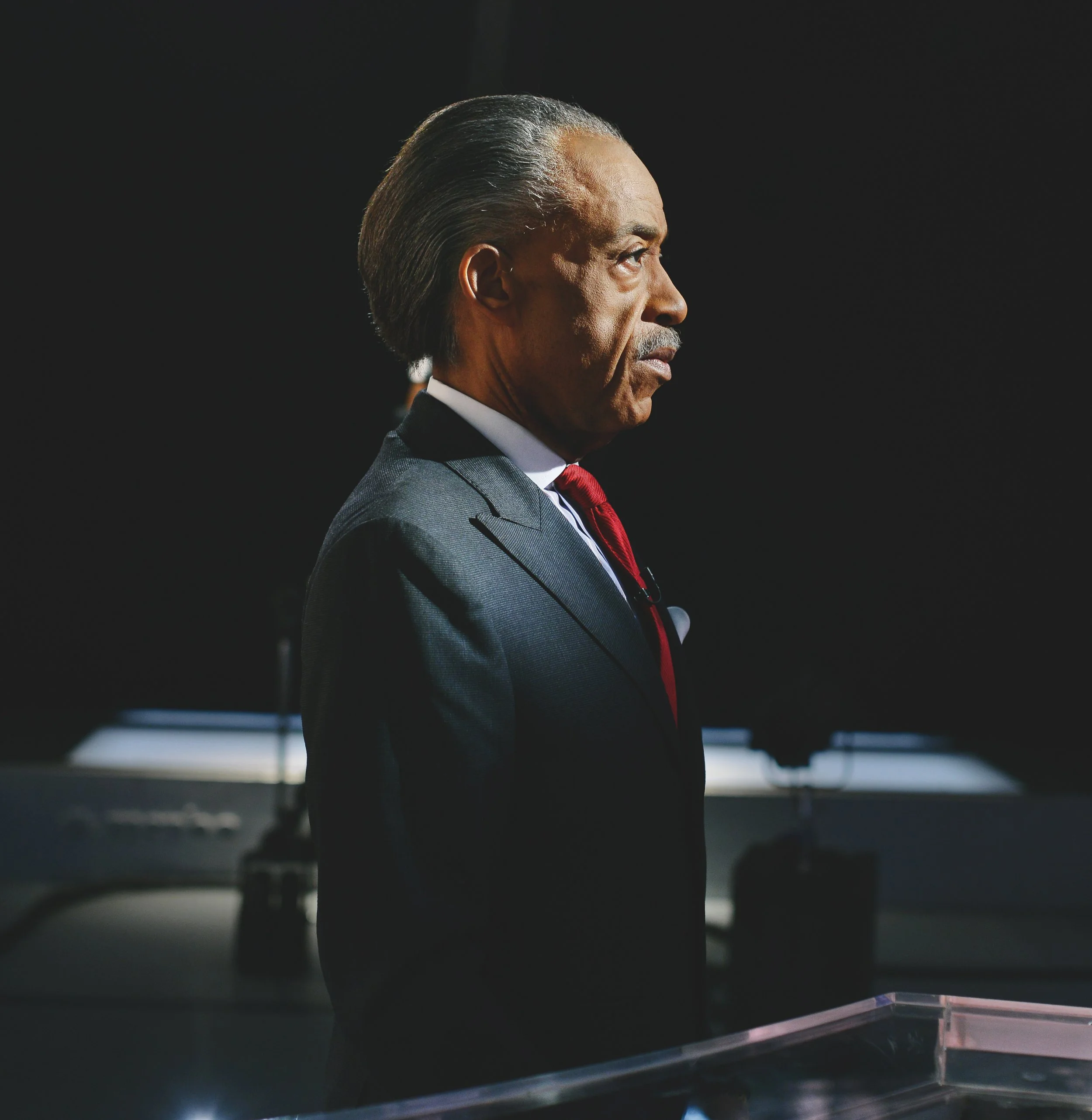 Side profile of an older man with slicked-back gray hair, wearing a dark suit, white shirt, and red tie, standing at a podium in a dark room.