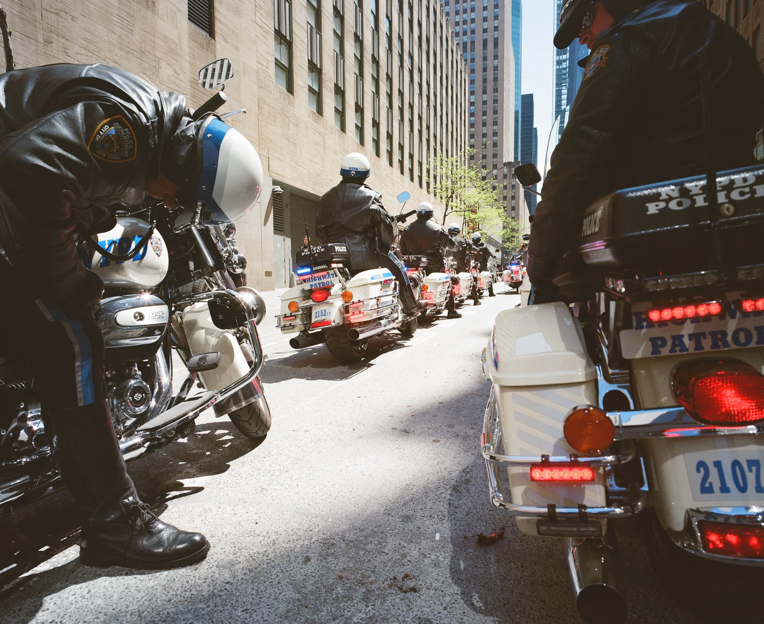 Line of police officers on motorcycles parked along a city street, with tall buildings in the background. The officers are dressed in black leather jackets and helmets, with some looking down or away from the camera. The street appears to be cleared 