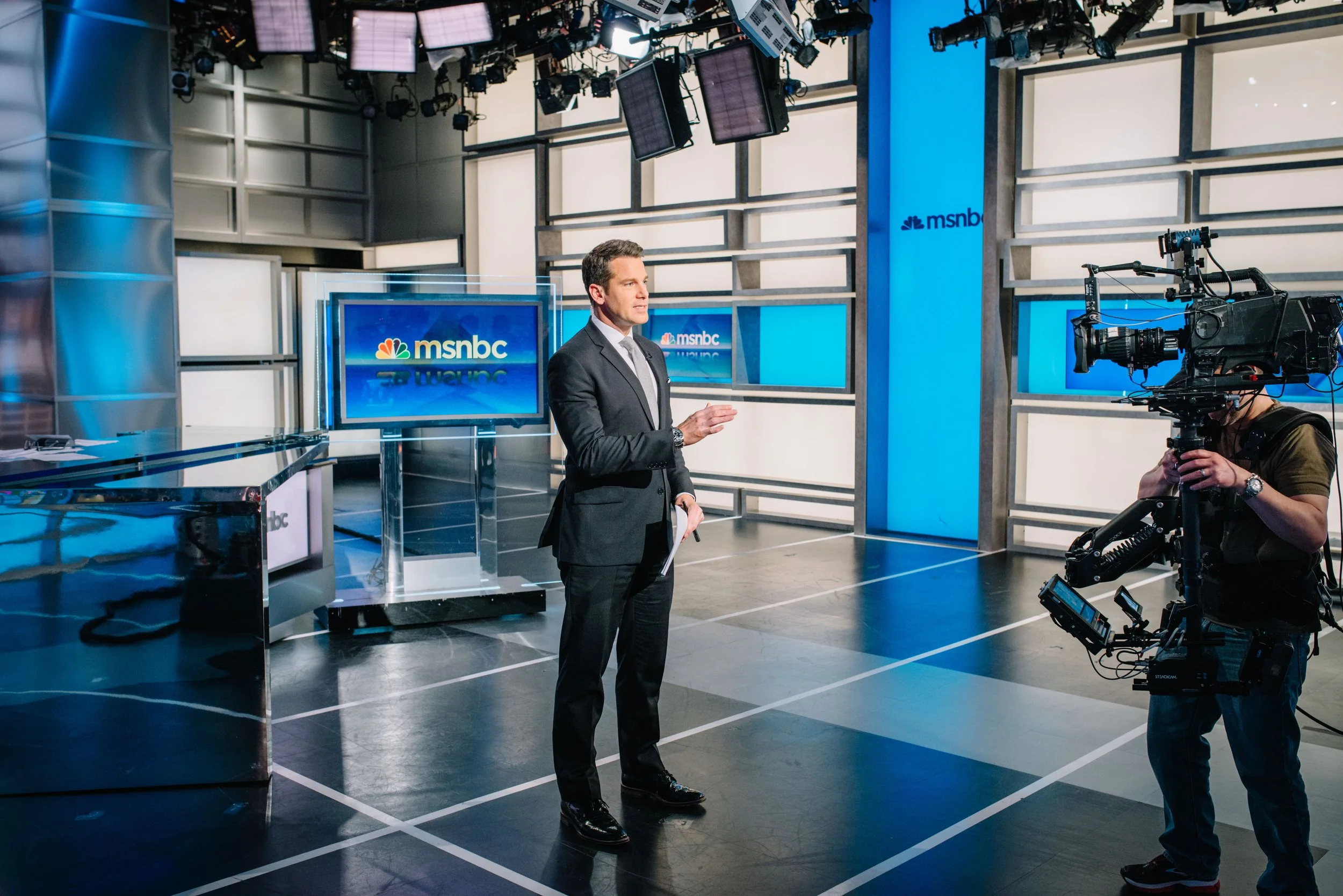 A man in a suit standing and speaking in a television studio with multiple MSNBC screens, a large camera on its operator, and professional lighting overhead.