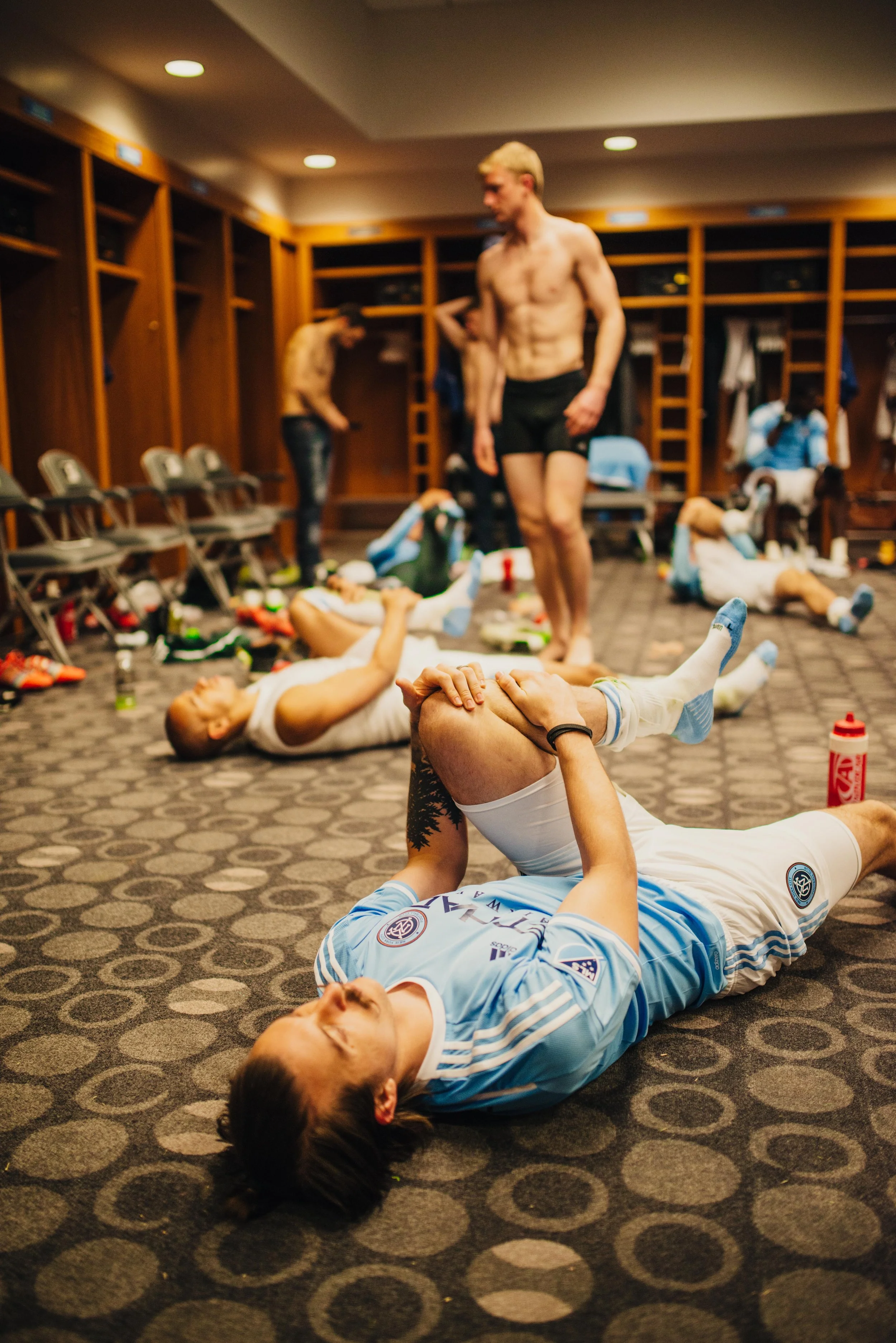 Soccer players in a locker room stretching and relaxing after a game, some lying on the floor and others standing.