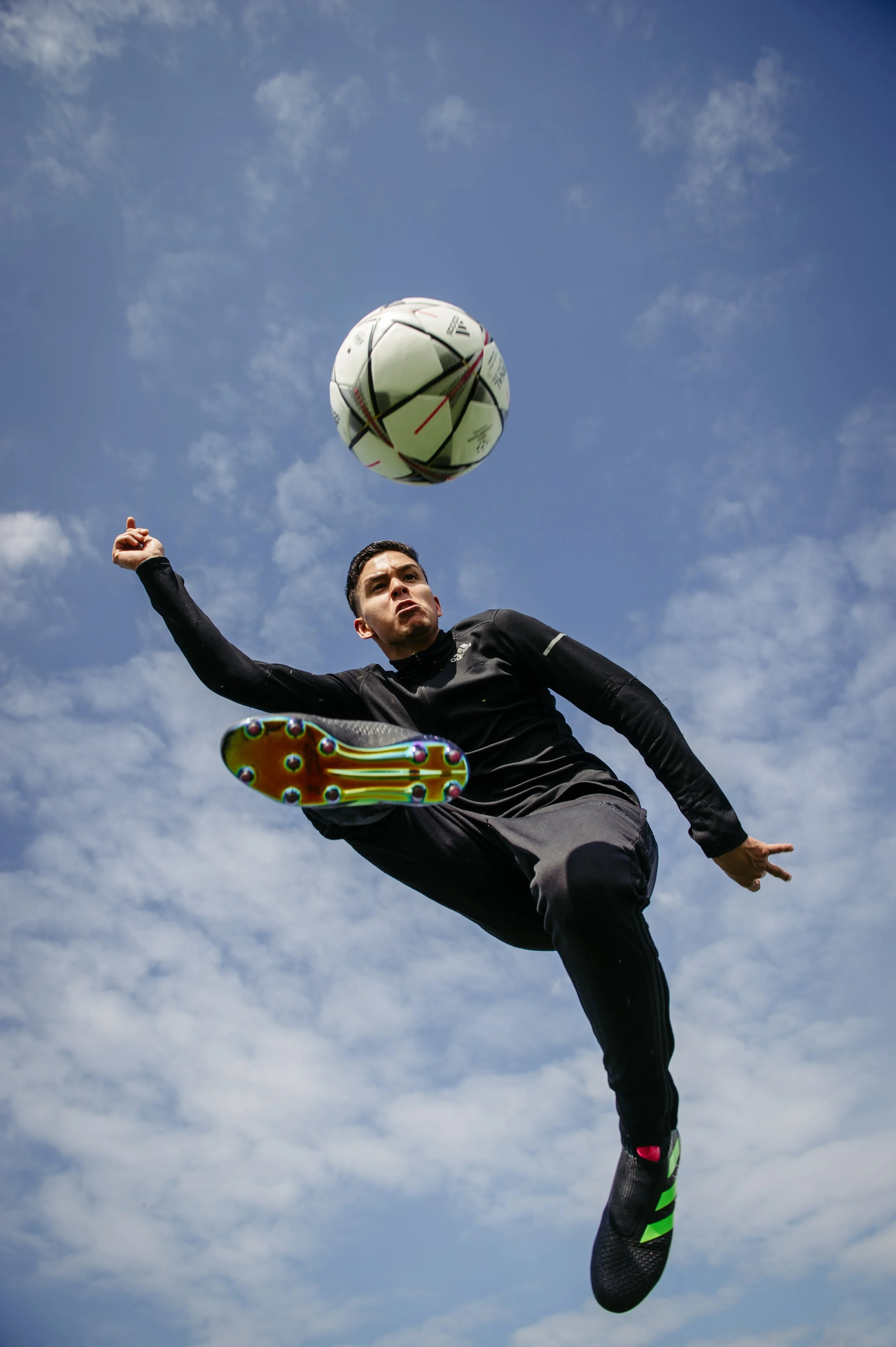 A soccer player in black athletic clothing executing a bicycle kick in mid-air during daytime with a partly cloudy sky.