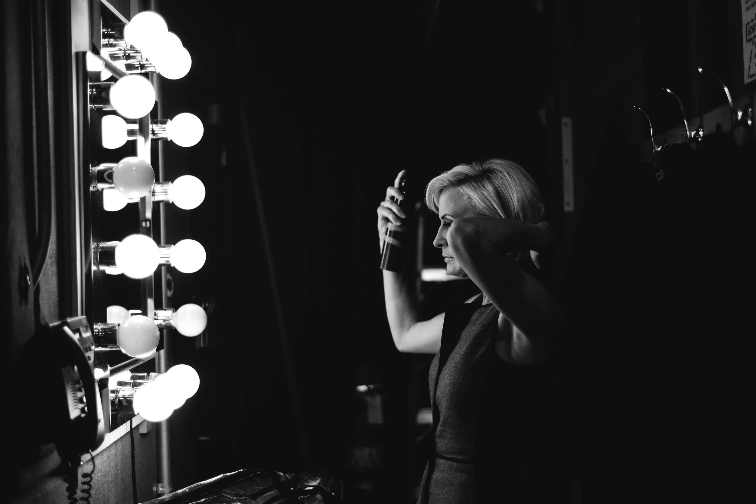 A woman with short blonde hair fixing her hair while standing in front of a mirror with bright round vanity lights, in a dressing room or backstage area.