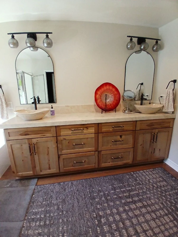 Bathroom vanity with two vessel sinks, large mirrors, and modern light fixtures; wooden cabinet, decorative red plate, and rug on the floor.