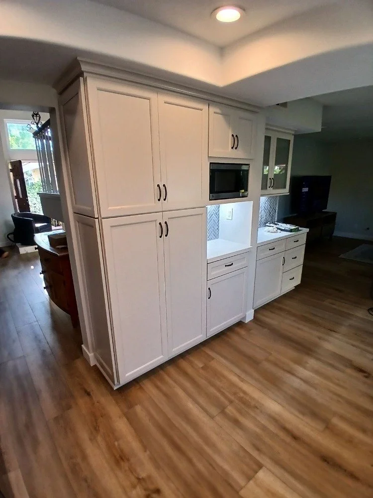 White kitchen cabinets with a microwave built into the cabinetry, situated in a room with wooden floors and adjacent to a living space with a TV.