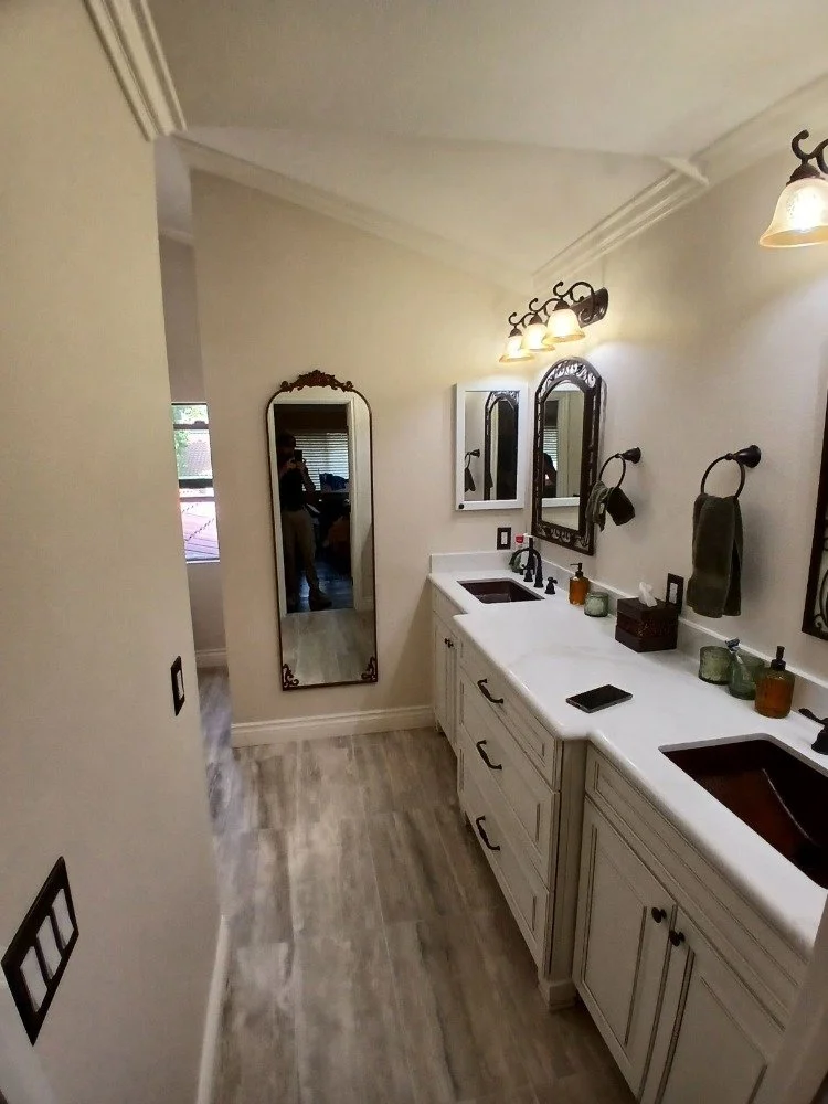 Bathroom with a double vanity, two brown sinks, three mirrors, and wall-mounted light fixtures. There are decorative soap dispensers and a box on the counter, with a full-length mirror on the left wall.