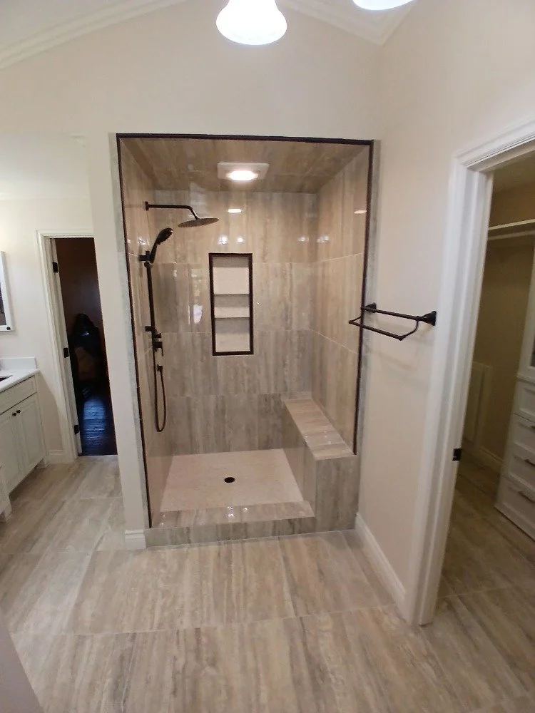 A walk-in shower with beige marble tiles, a built-in niche, a rain showerhead, and a bench, enclosed by glass walls in a bathroom.