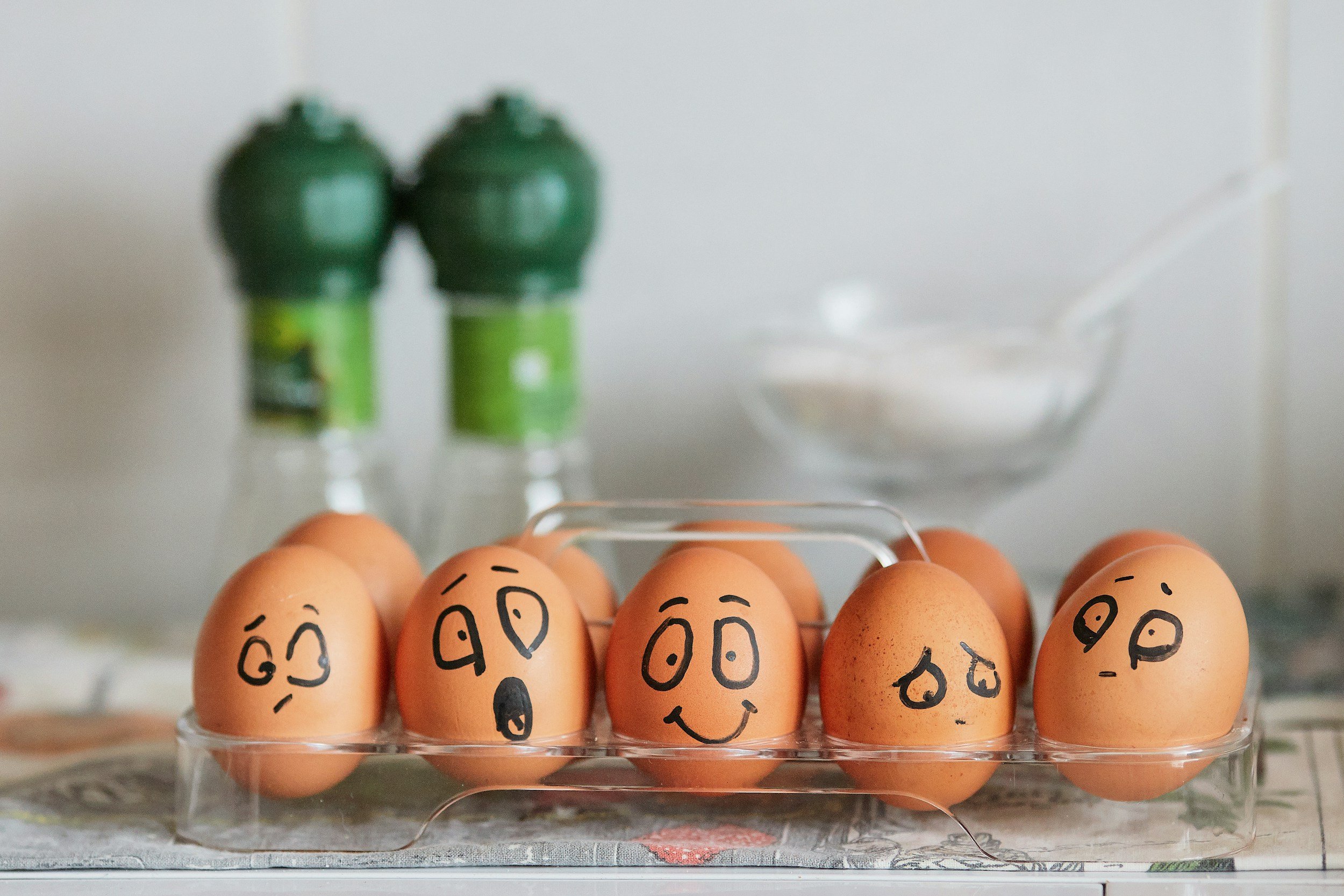 Eggs with hand-drawn sad and happy faces arranged in a carton on a kitchen counter.