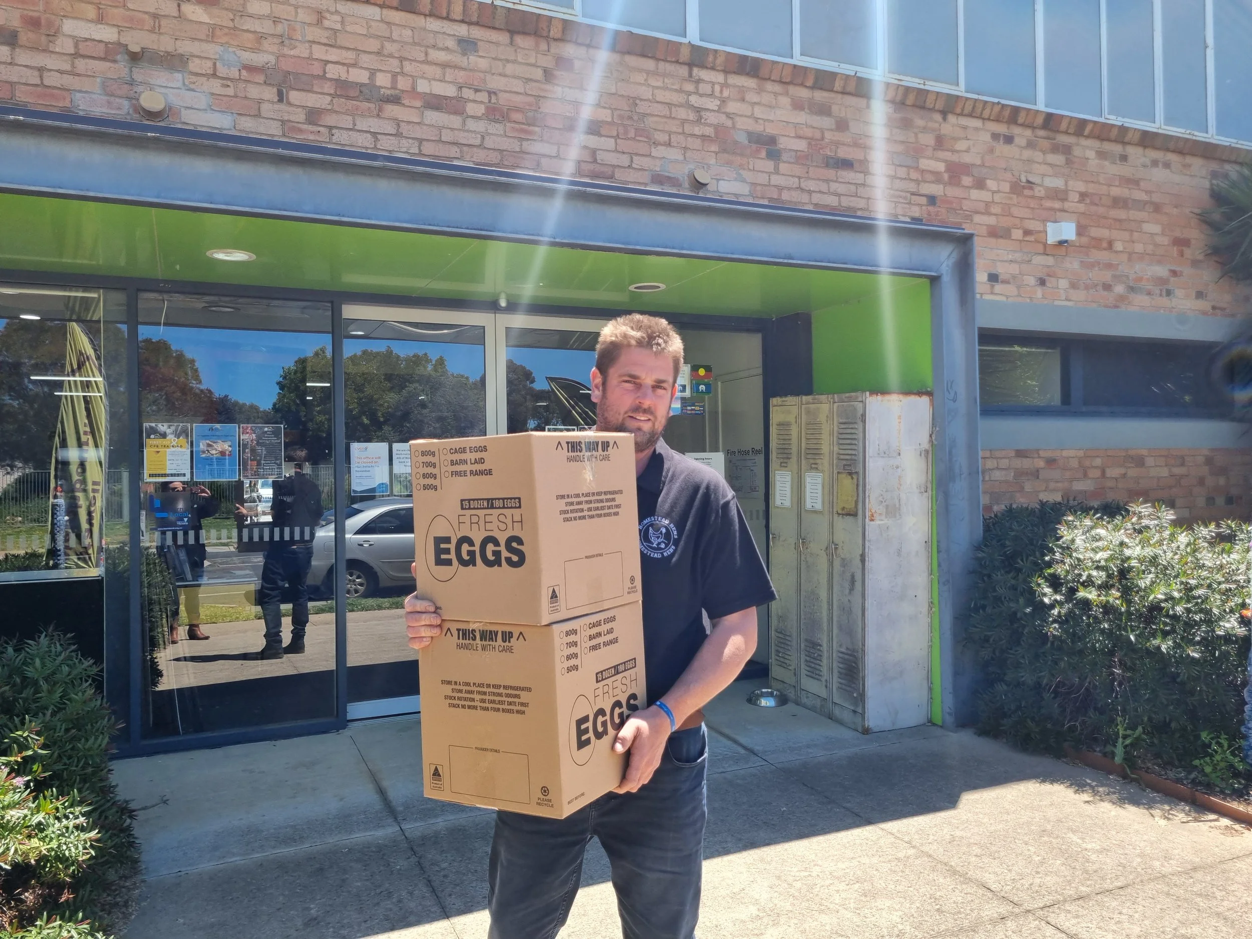 Josh Pitt from Homestead Hens holding a large box of fresh eggs outside a building with glass doors and locker storage in the background for their Egg Fundraiser.